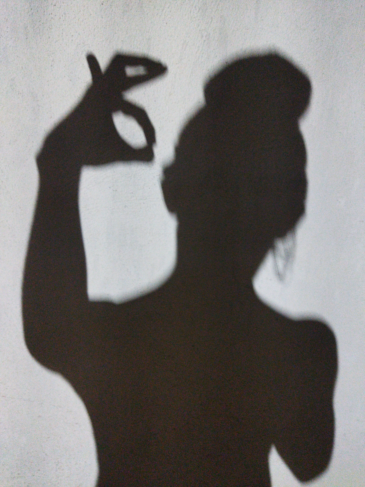 A stark shadow of a person is cast against a light-colored wall. The person is facing away from the viewer, their hair gathered in a bun on top of their head. Their right arm is raised, and their hand is making an "OK" gesture, with the thumb and index finger forming a circle. The shadow suggests a partial view of the person's torso and shoulders. No information is apparent from the image to indicate its location as Tinaquillo, Venezuela.
