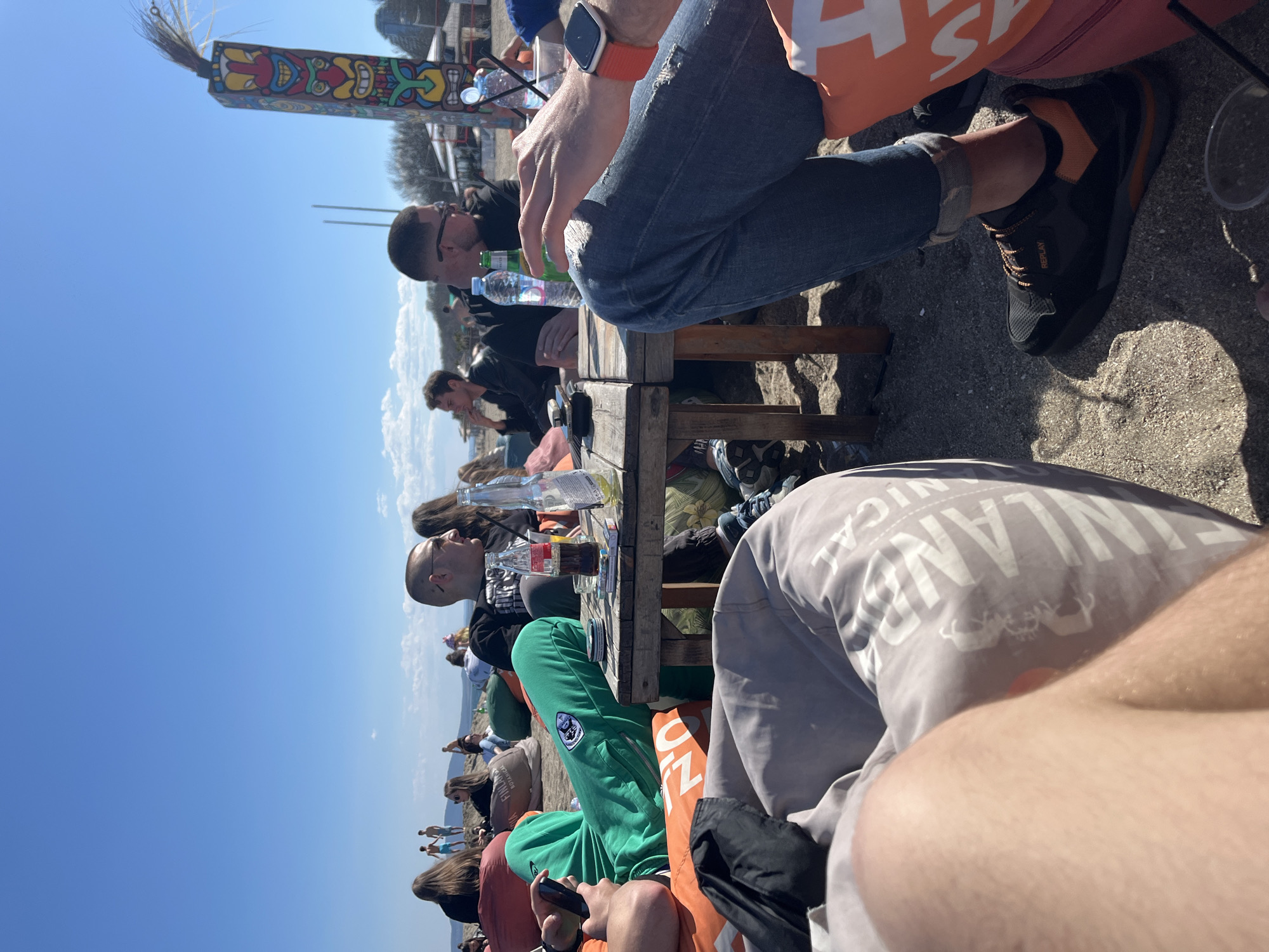 A group of people are seated at a wooden table on a beach, possibly a cafe or restaurant setting. They appear to be relaxing and socializing in Burgas, Bulgaria. A totem pole is visible in the background. Some individuals are drinking from bottles and glasses. A beach and other people are near the water's edge. One person is wearing shoes with Replay branding.
