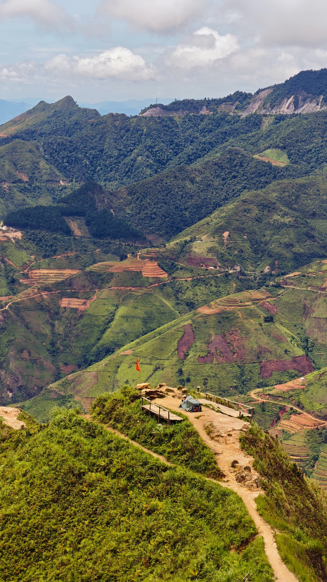 The image captures a breathtaking panoramic view of the mountainous landscape of Bắc Yên, Vietnam, characterized by its dramatic terraced fields. In the foreground, a winding dirt path leads to a small, elevated viewpoint. At the viewpoint, a wooden platform with railings has been constructed, offering a clear vantage point. A red flag stands planted on the rocky ground to the left of the platform. A green tent is set up on the platform, with what appears to be a person or belongings inside.
The midground and background are dominated by lush green mountains, intricately carved with vibrant orange and red terraced fields, evidence of agricultural activity. These terraces create a striking visual pattern against the natural contours of the hills. Roads and pathways snake through the terrain, connecting the different levels of the terraces. The sky above is partially filled with white, fluffy clouds, casting soft shadows across the dramatic landscape. The overall impression is one of a remote and scenic location, showcasing the beauty of rural Vietnam and the ingenuity of its people in cultivating the challenging mountainous terrain.
