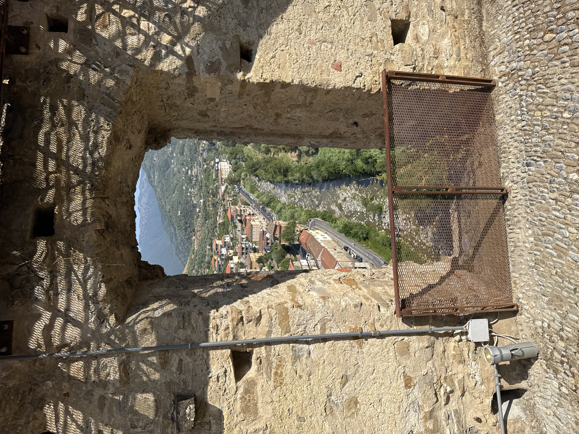 The image captures a view through an arched stone opening in a building, looking out onto the town of Dolceacqua, Italy. The opening reveals a valley with a river, lined by trees and a road with cars. Buildings with terracotta roofs are clustered on the hillsides. The foreground consists of a cobblestone path and a rusted metal grate. The stone walls of the building framing the view are textured and show signs of age. Sunlight casts shadows through a patterned structure above the archway, creating a textured effect on the stone. Electrical conduits and a light fixture are visible on the left side of the stone wall. The scene is bathed in daylight, indicating it was taken during the daytime. There are no people visible in the image.