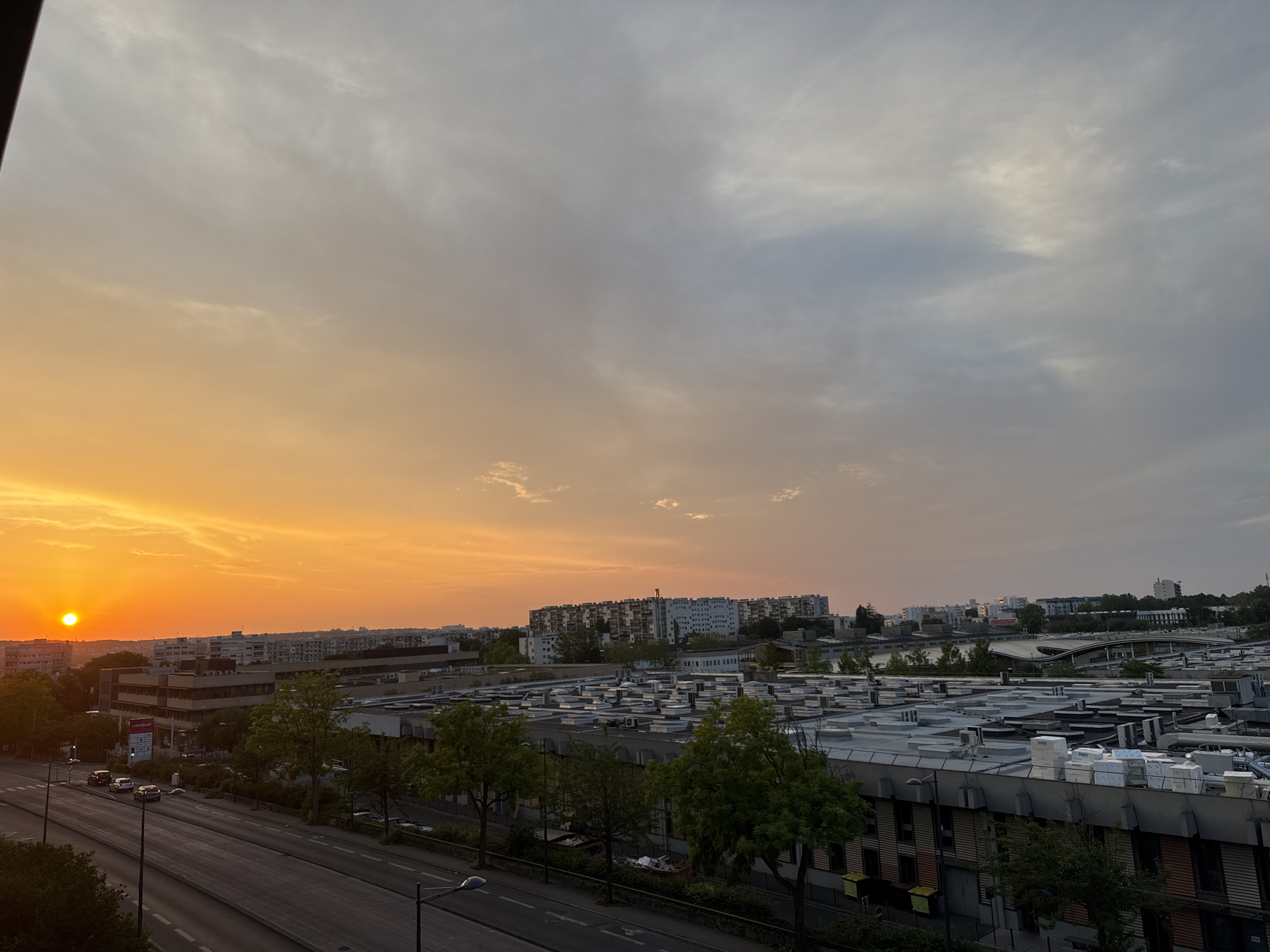 The image captures a wide-angle view of an urban landscape at sunset. The left side of the frame is dominated by the vibrant orange glow of the setting sun, positioned low on the horizon, casting a warm light across the sky. Clouds are present, with the sky transitioning from a bright orange near the sun to a more diffused grey and blue towards the right.
In the mid-ground, a series of buildings are visible. Closest to the viewer is a large, flat-roofed industrial or commercial building, characterized by numerous HVAC units and piping on its roof. This building has a dark facade with visible windows. To the left of this structure, a multi-story office building with a lighter facade and darker window frames is present, with signage visible on its exterior. Beyond these, a line of residential apartment buildings stretches across the horizon, appearing as a dense collection of structures.
In the foreground, a multi-lane road runs horizontally across the lower portion of the image. Several cars are visible on the road, appearing as dark shapes. Streetlights and trees line the road. The overall scene depicts a transitional period of the day in a developed urban area, with a mix of residential, commercial, and industrial elements.
