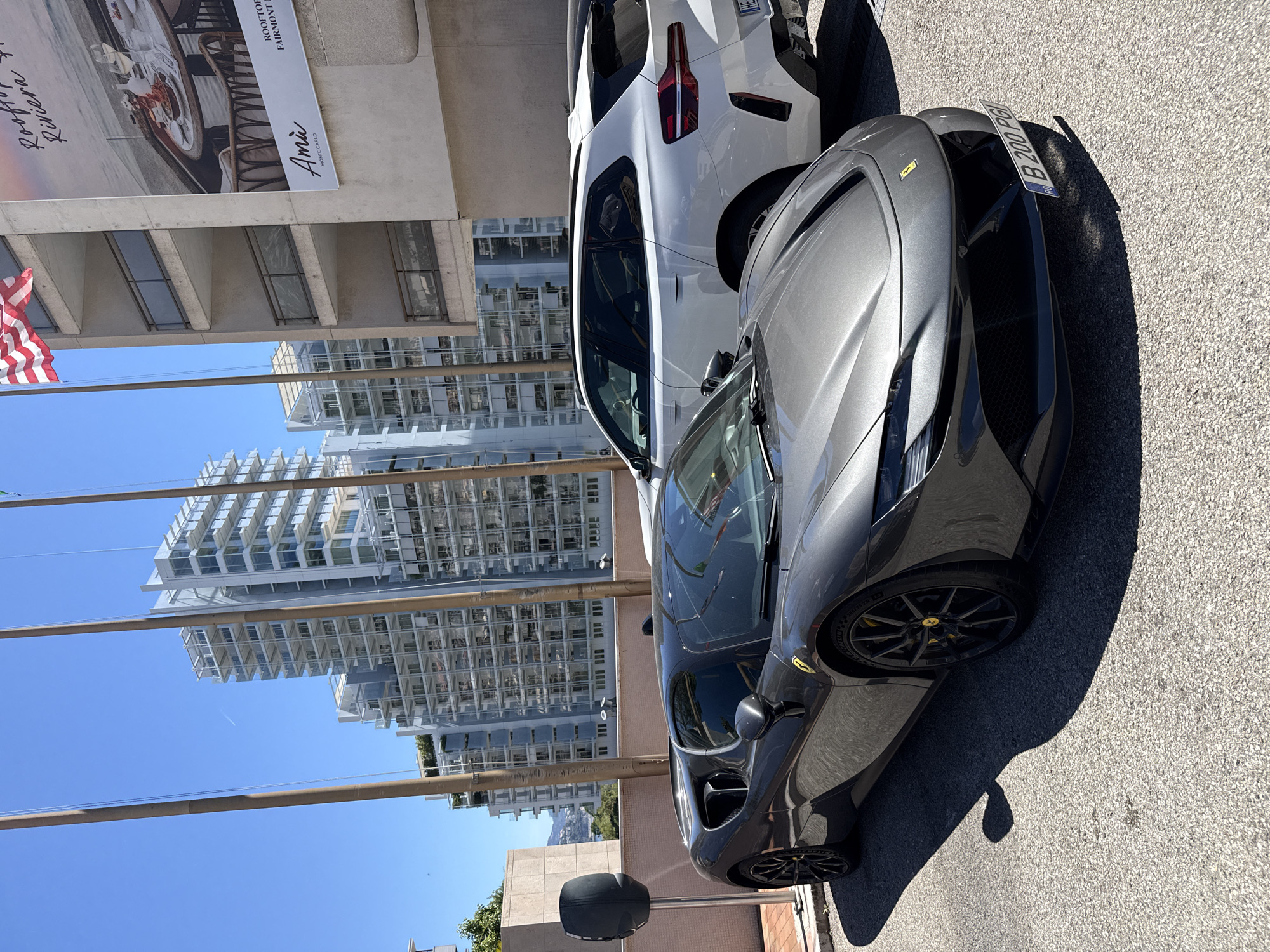 In front of a modern building with multiple balconies, a dark grey Ferrari sports car is parked on an asphalt surface. To the right of the Ferrari, a white BMW SUV is also parked. In the background, several tall, contemporary buildings with glass facades rise against a clear blue sky. A billboard featuring a table setting and the text "Amià Monte Carlo" is visible on the right side of the building. The scene appears to be a sunny day in a city setting, likely Monaco, given the presence of luxury vehicles and upscale architecture. An electric car charging station is also visible to the left of the Ferrari.