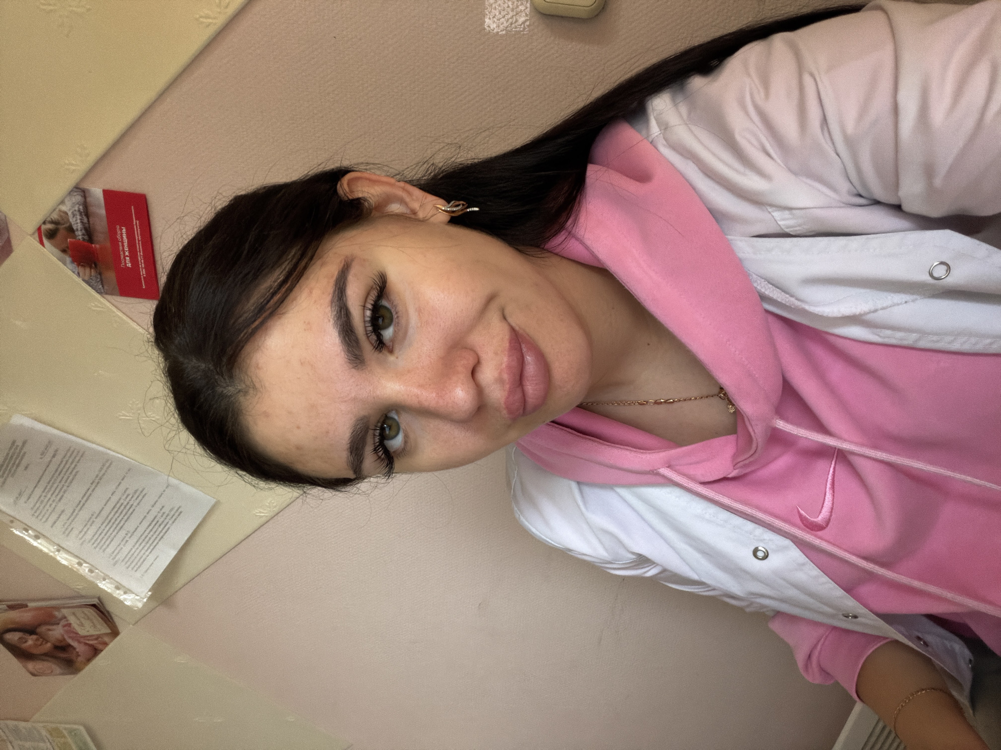The image shows a medium-close-up, indoor portrait of a young woman with dark hair, light skin, and green eyes. She is wearing a pink hooded sweatshirt with a Nike logo and a white jacket over it. She has on makeup, including false eyelashes, and is wearing gold earrings and a gold necklace. The background consists of a pink-colored wall with some papers and a red advertisement affixed to it. The lighting suggests an artificial indoor environment. There is no information in the image to determine the location within the city of Garevaya, Russia.