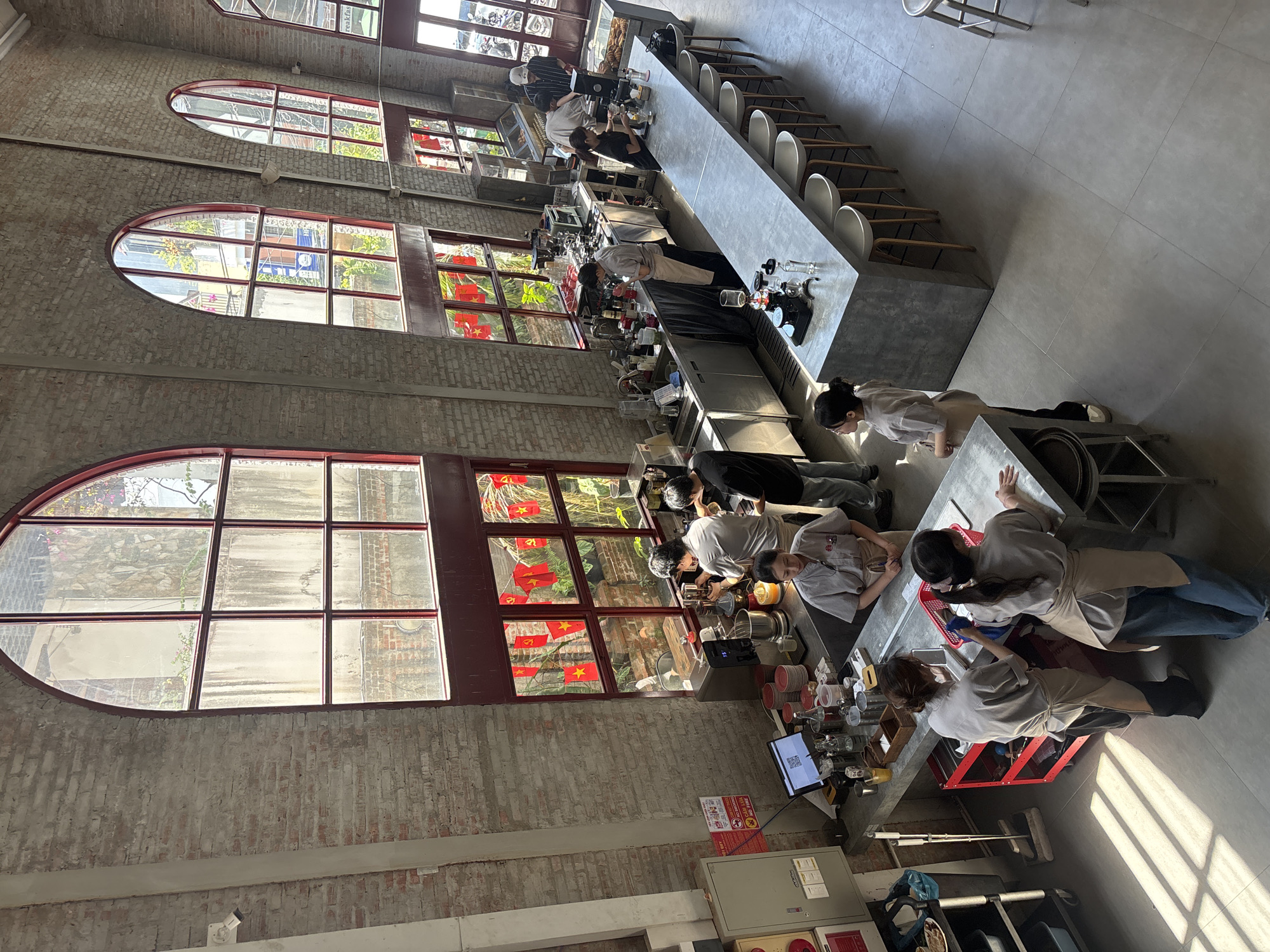 This is an aerial, wide-angle shot of a busy cafe in Đà Nẵng, Vietnam, featuring an industrial aesthetic with exposed brick walls and large, arched windows. Sunlight streams in, casting distinct shadows across the tiled floor.
In the background, several people are visible. On the right, a group of patrons are seated at a long, concrete bar. Behind the bar, cafe staff are working. One person in a dark shirt and cap is interacting with a customer at the far end of the counter, near a display of pastries. Another person in a white shirt and apron is preparing drinks behind the counter, using a coffee machine.
Towards the center and left of the image, more cafe staff are engaged in various tasks. Three individuals in grey shirts and aprons are behind a counter, with one appearing to operate a coffee machine and another attending to drinks. Two other individuals are on the other side of the counter, facing away from the camera, seemingly customers or staff members interacting with each other. A metal cart with various items is positioned to the left of the counter.
The cafe's interior is characterized by raw brick walls, concrete countertops, and large windows with red frames. Red and yellow banners are hanging in some of the windows. Various pieces of cafe equipment, including coffee machines, blenders, and shelves stocked with cups and bottles, are visible. The overall atmosphere appears to be bustling and well-lit.
