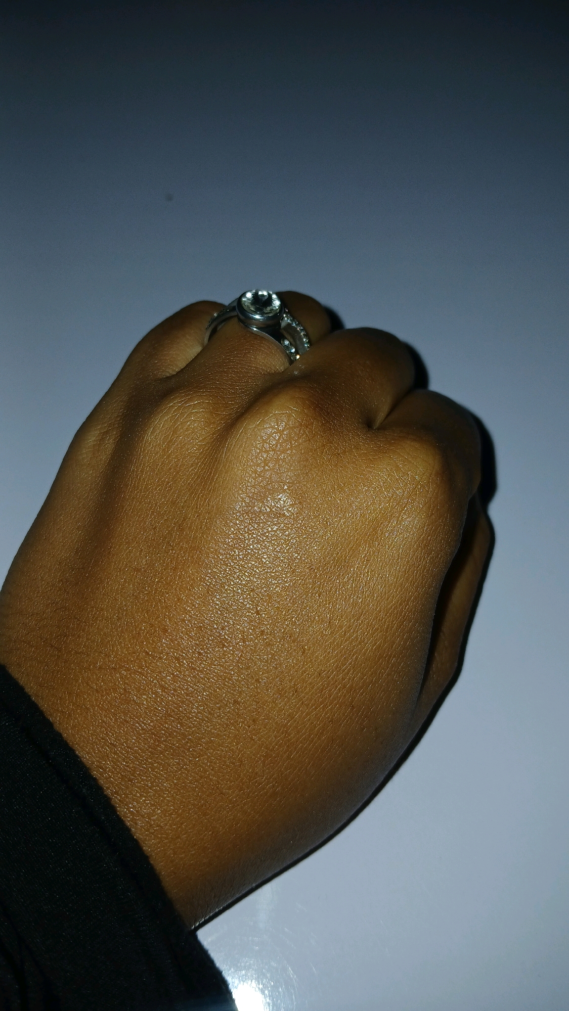 A close-up, low-angle shot shows a person's hand, wearing a silver ring with a clear stone, against a blurred background that appears to be a plain wall or surface. The hand is held in a somewhat closed fist, with the knuckles and finger joints clearly defined, and the skin is a warm brown tone. The ring is positioned on the index finger, and its band is also adorned with small, sparkling stones. The lighting suggests a dimly lit environment, with a spotlight illuminating the hand and ring. The image does not provide any specific details about the location in Gura Top, Nigeria.