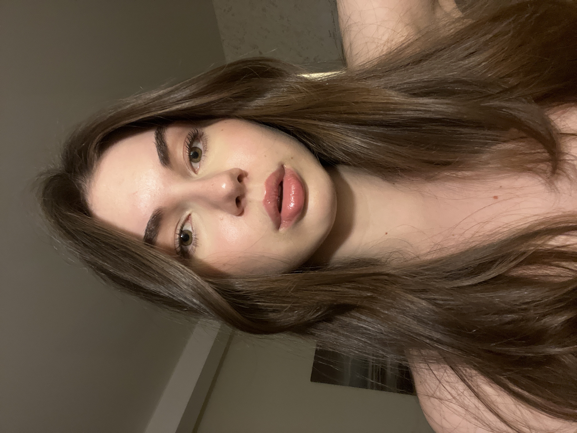 A portrait-style photograph features a young woman with light skin and brown hair. Her hair is long, glossy, and styled with a subtle wave. Her facial features include light-colored eyes, full lips with a glossy finish, and defined eyebrows. The photograph appears to be taken indoors, with an ambient light source. The background is a plain wall, suggesting a domestic setting. The overall composition is simple and focuses on the subject's face and hair. There is no indication of location of St. Petersburg, Russia.