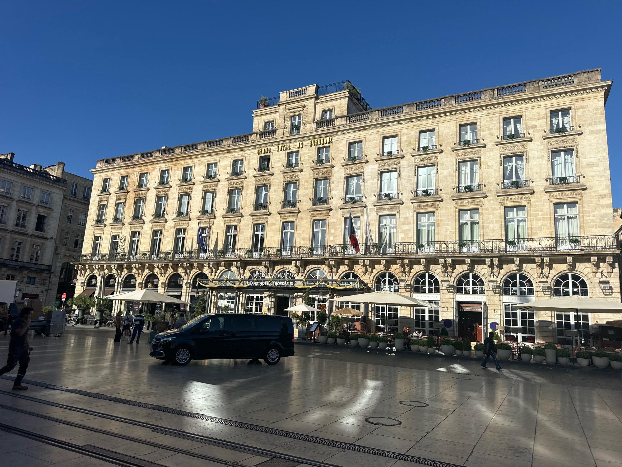 The image captures a sunny day in Bordeaux, France, specifically in front of the InterContinental Bordeaux - Grand Hotel de Bordeaux. A large, ornate building with classical architecture dominates the background. The facade is made of light-colored stone, featuring multiple rows of windows with decorative surrounds and balconies. The building has a prominent sign that reads "INTERCONTINENTAL GRAND HOTEL DE BORDEAUX."
In the foreground, a wide, paved plaza with tram tracks stretches across the scene. A dark-colored van is parked on the right side of the plaza, and several people are visible walking and sitting at outdoor tables beneath umbrellas, suggesting a cafe or restaurant area associated with the hotel. The sky is clear and blue. Sunlight casts long shadows across the plaza, indicating it is likely late morning or early afternoon. The overall scene depicts a bustling urban environment with a mix of grand architecture and public space.