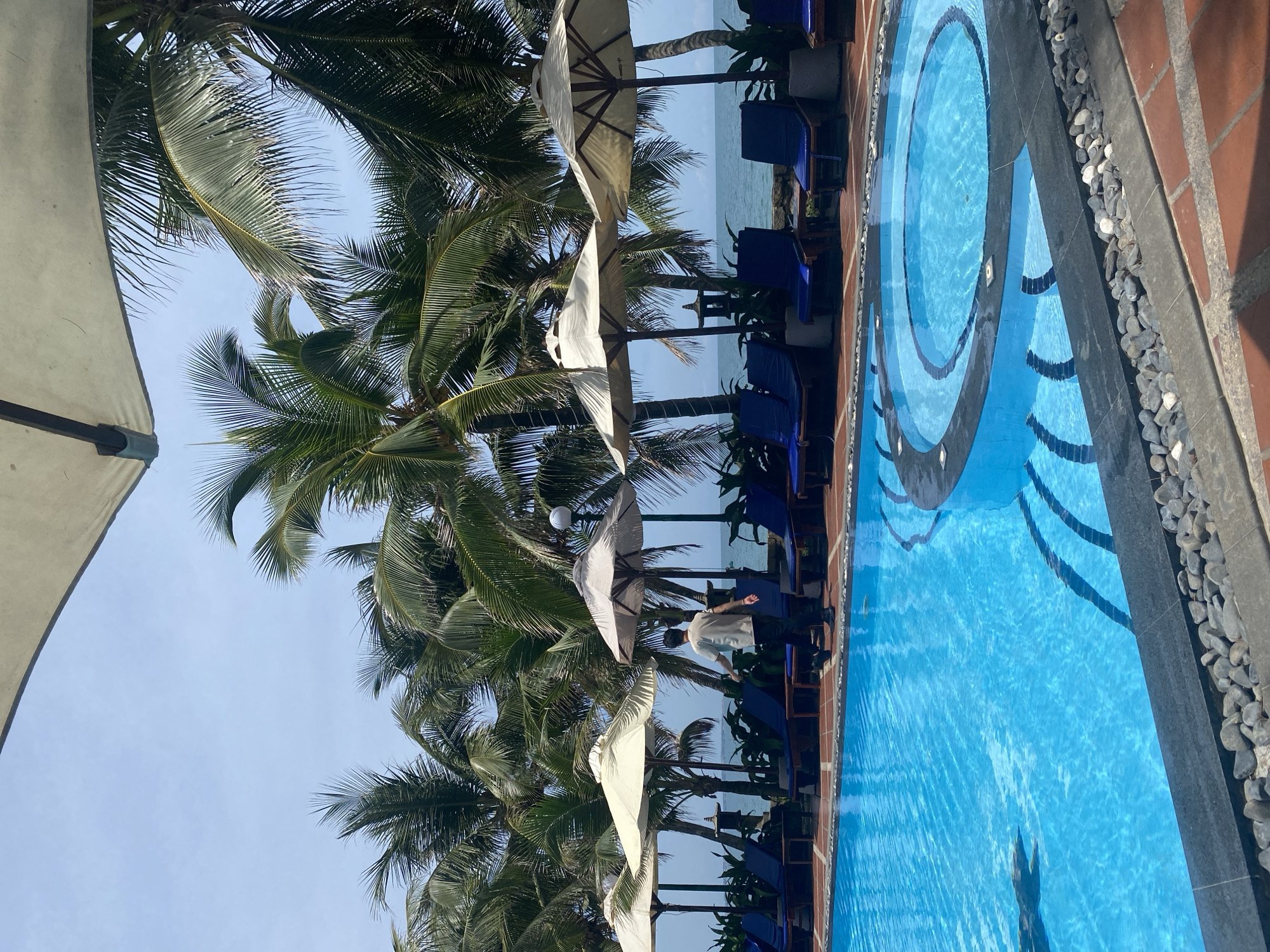 The image depicts a resort swimming pool area in Phu Thuy Ward, Vietnam. The pool is bright blue and features a circular hot tub within its expanse. Surrounding the pool are several lounge chairs with blue cushions, partially shaded by cream-colored umbrellas. Palm trees line the background, with the ocean visible beyond them. A lone individual, dressed in a light-colored t-shirt and dark pants, walks away from the camera near the lounge chairs, suggesting a relaxed, resort-like atmosphere. The scene is set under a clear blue sky.