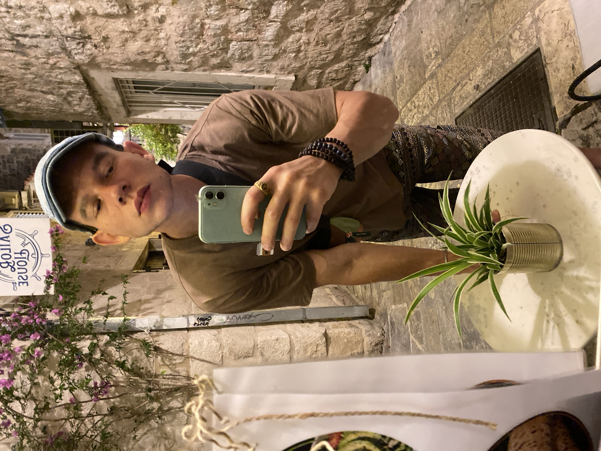 The image shows a young person taking a mirror selfie in an alleyway in Budva, Montenegro. The person is wearing a brown t-shirt, patterned pants, and a blue cap. They have several bracelets on their left wrist and a ring on their right index finger. In the foreground, a small plant sits in a tin can on a white table. Behind the person, a stone building with a barred window and a flowering vine is visible. The alleyway is paved with cobblestones. A sign with Cyrillic script is partially visible in the background. The lighting suggests it is evening or nighttime.