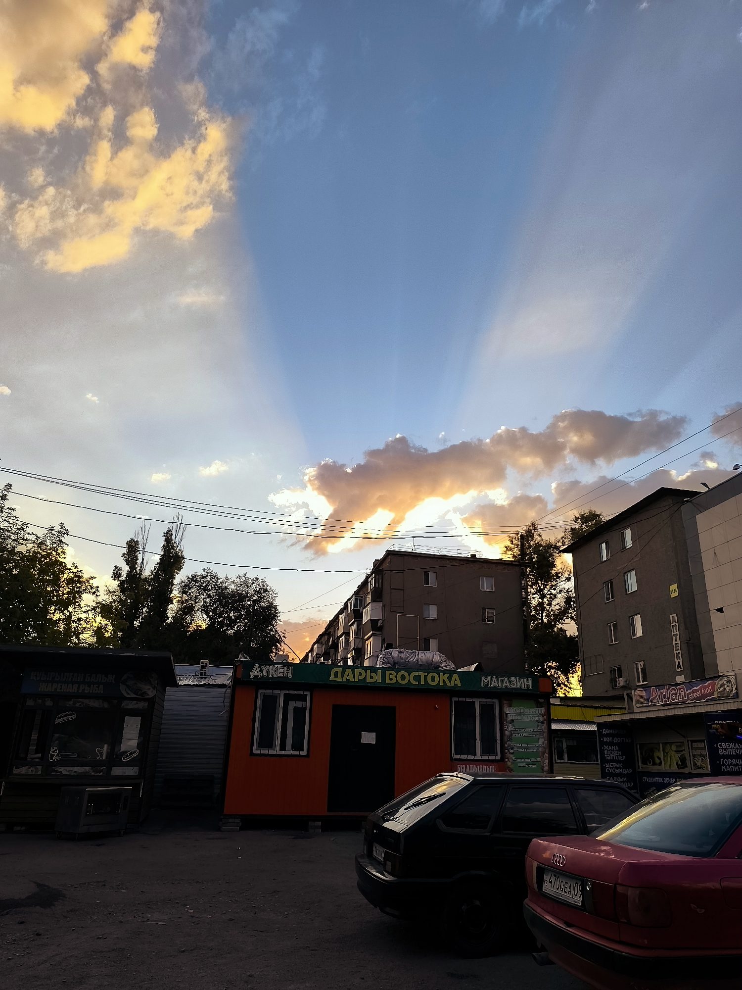 A view of buildings and a street scene at dusk in Qarağandı, Kazakhstan, with dramatic crepuscular rays piercing through the clouds. In the foreground, a paved area shows two parked cars; a dark-colored hatchback on the left and a red sedan on the right. Behind the cars, there are small commercial structures, one painted orange with "АУКЕН" and "ДАРЫ ВОСТОКА" visible on its facade, and another to its left with signage indicating "КУЫРЫЛГАН БАЛЫК" and "ЖАРЕНАЯ РЫБА." Further back, two multi-story residential buildings are visible, one darker gray and the other lighter gray, with windows and balconies. Trees with green foliage are scattered throughout the scene, silhouetted against the colorful sky. Power lines crisscross the upper portion of the image. The sky itself is a mix of deep blue and warm, golden hues where the clouds are illuminated by the setting sun, creating prominent rays of light. The overall atmosphere is serene yet striking due to the celestial display.