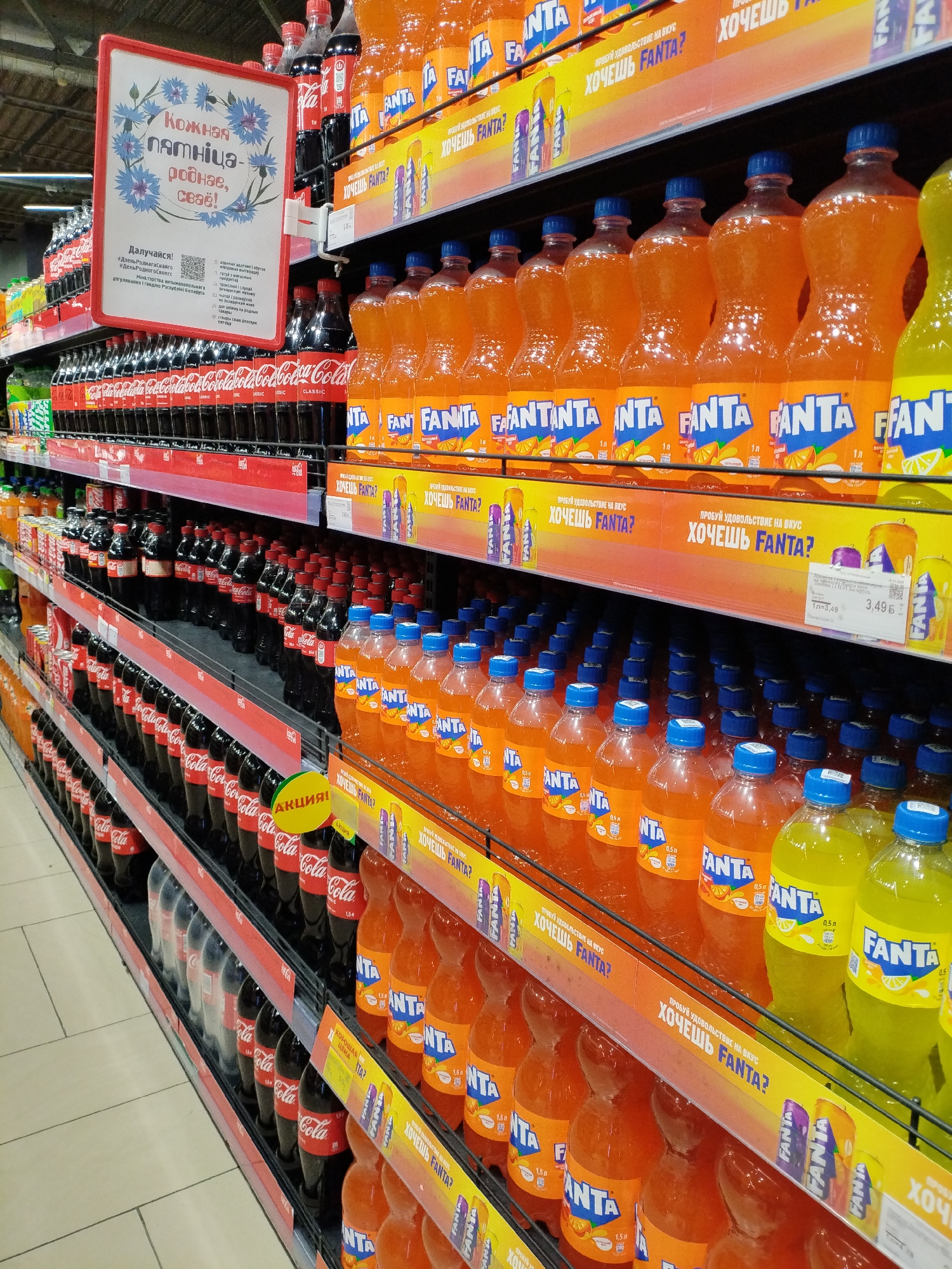 The image depicts a retail environment, likely a supermarket aisle, showcasing a wide selection of beverages. The primary focus is on rows of plastic bottles containing dark brown and orange-colored sodas, prominently displaying brands like Coca-Cola and Fanta. Many bottles are arranged on shelves, some are stacked, and a few are in multipacks. Yellow promotional banners with Russian text, including "Хочешь Fanta?" (Do you want Fanta?), are visible above the Fanta bottles. A white sign with blue floral illustrations and Cyrillic text hangs from the top shelf, seemingly a promotional or informational display. The store features light-colored tiled flooring and metal shelving units. The scene suggests a typical beverage section in a store, with ample stock and clear product display. The specific location of Mahilyow, Belarus, is not visually identifiable within the provided image; it is external information.