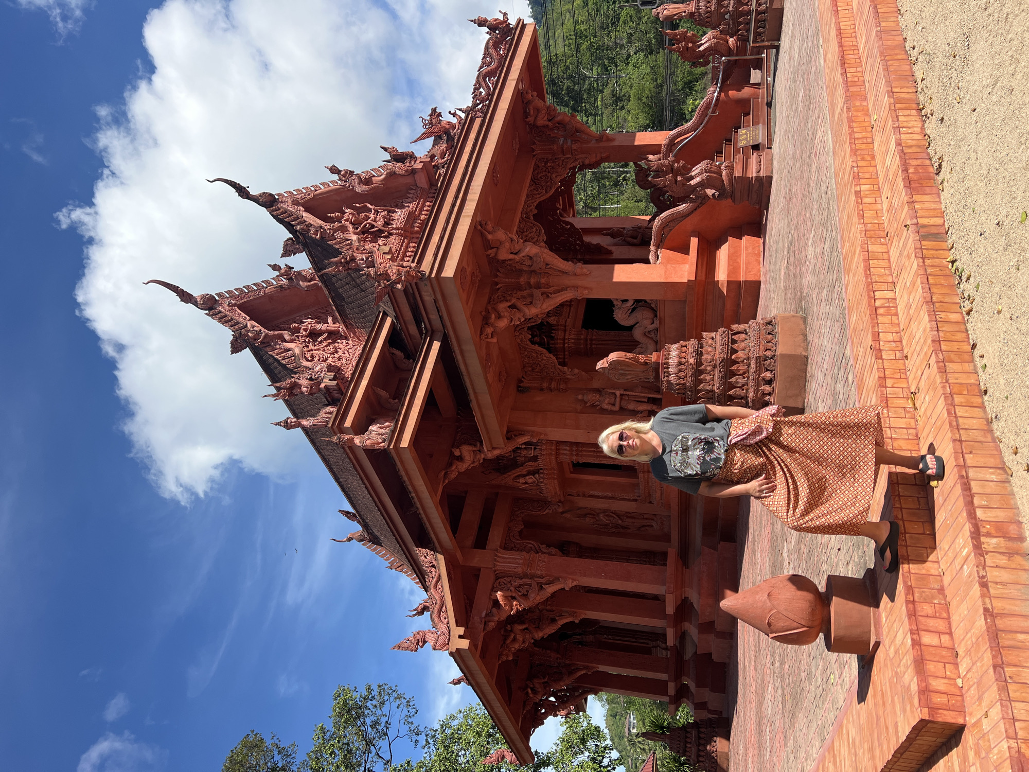 A woman stands on the steps of a vibrant orange temple. She is wearing sunglasses, a dark t-shirt with a graphic print, and a patterned skirt. Her hair is blonde and shoulder-length. She is positioned in front of the temple entrance, which is adorned with intricate carvings and dragon motifs. The temple itself is a striking orange color, with multiple tiered roofs and ornate architectural details. The ground in front of the temple is paved with orange bricks, and a decorative lotus-shaped sculpture is visible to the left of the woman. The background shows lush green trees and a bright blue sky with scattered white clouds. The scene appears to be in a warm, tropical climate, consistent with the location of Baan Hua Thanon, Thailand.