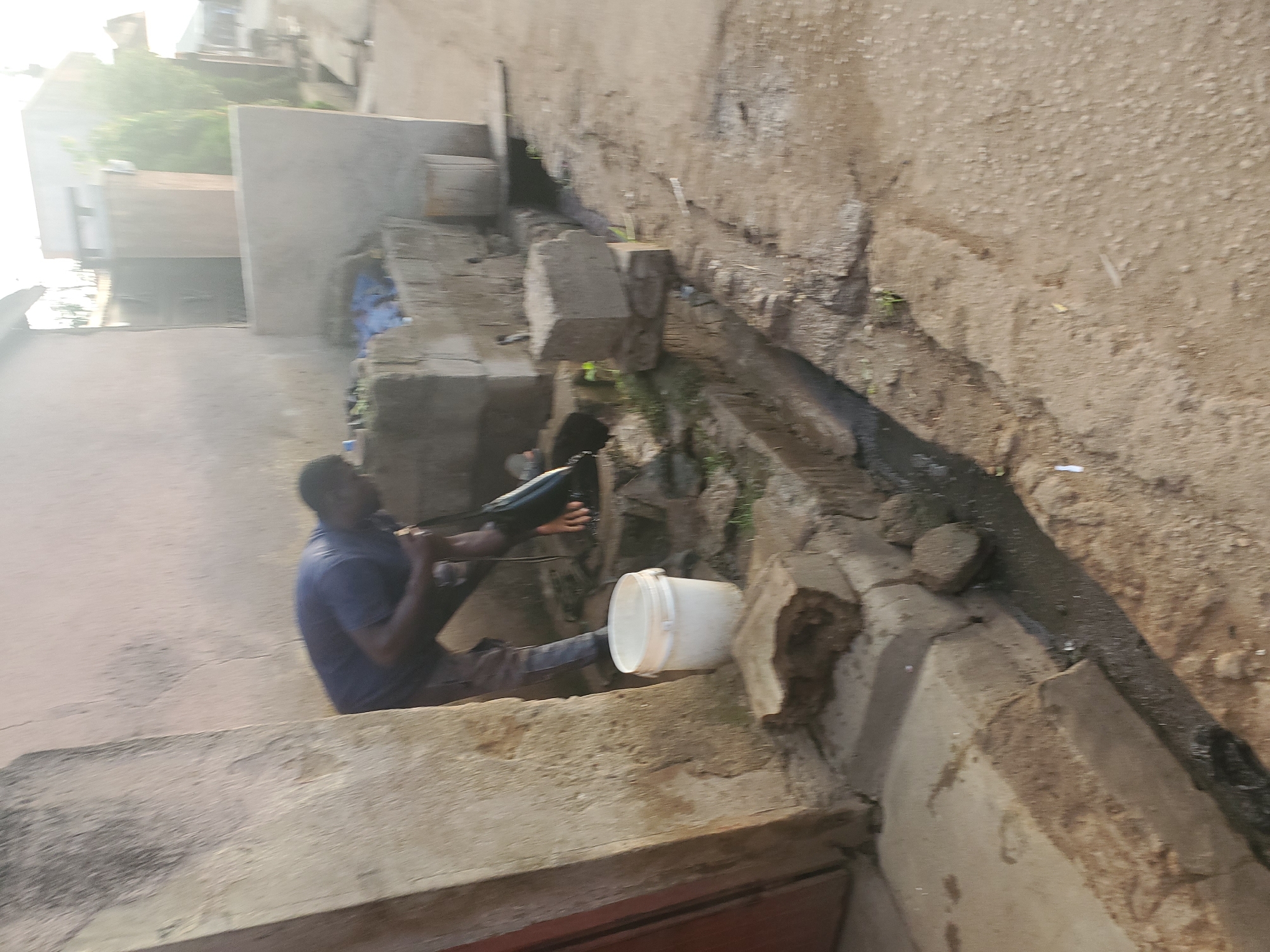 A man wearing a blue shirt and grey pants is standing next to a concrete building in what appears to be an alleyway or side street in Jos, Nigeria. He is bent over, holding a black bag or object and appears to be interacting with a white bucket. The area is somewhat dilapidated, with cracked concrete, visible debris, and an open drainage channel running along the ground. The overall scene depicts a typical urban environment with signs of wear and tear.