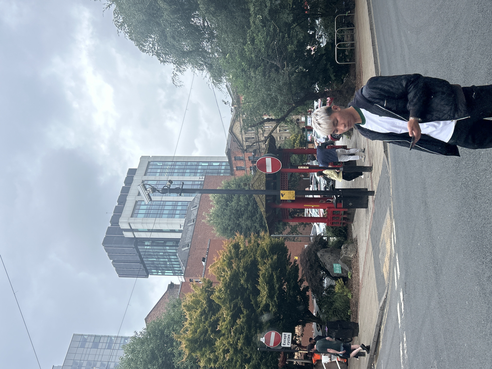 The image depicts a street scene in Manchester, United Kingdom, likely within the city center or a commercial district, given the presence of modern high-rise buildings and an urban street layout. The sky is overcast, suggesting a cloudy day.
In the foreground, a young man with light-colored hair is walking on the street, looking down at his smartphone. He is wearing a black quilted jacket over a white t-shirt and dark trousers. To his left, a street corner features a pedestrian crossing and a road with parked cars visible in the background.
Further back, a traditional-looking red pavilion with a tiered roof, reminiscent of East Asian architecture, stands prominently. Adjacent to it is a traffic sign indicating "Except cycles." Behind the pavilion, several trees with green foliage are scattered throughout the scene.
In the background, several buildings of varying architectural styles are visible. A tall, modern glass-fronted skyscraper dominates the left side of the frame, juxtaposed with older brick buildings and another contemporary building with a dark facade. A security camera is mounted on a pole near the red pavilion. Several people are walking in the middle ground, and more vehicles are parked or driving in the background. The overall atmosphere is that of a typical urban environment on a slightly grey day.