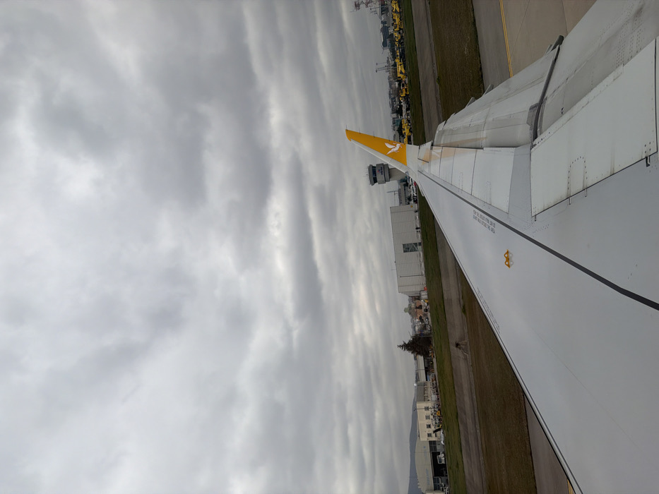 The image depicts an airport scene from the perspective of an aircraft window, showing a portion of the right wing in the foreground. The location is Sabiha Gökçen International Airport (ISG) in Pendik, Istanbul, adjacent to Tuzla, Turkey.
The aircraft wing, predominantly white with a dark gray leading edge and black stripe, extends from the lower right into the mid-ground. The winglet or vertical stabilizer, positioned towards the rear of the wing, is yellow and features the Pegasus Airlines logo (a stylized bird). Visible markings on the wing include text such as "NO STEP" and "DO NOT STEP ON THIS AREA" in black. The wing's flaps and slats appear retracted or partially extended.
Beyond the wing, an airport taxiway or tarmac is visible, characterized by gray asphalt and yellow painted lines. Adjacent to this, a strip of green grass and brown earth separates the immediate ground from airport buildings. In the mid-ground, a multi-story airport control tower stands prominent, gray in color, with a visible "ISG" logo near its top. To the left of the control tower, several industrial-style airport buildings and hangars are present. To the right of the control tower, numerous yellow ground support vehicles are parked in an organized row on the tarmac. A distant cityscape or hills are faintly visible on the horizon behind the airport infrastructure.
The sky is entirely overcast with a dense layer of gray clouds, indicating a cloudy or gloomy day. No discernible individuals or specific human actions are clearly depicted within the scene. The overall impression is one of an aircraft on the ground at an operational airport.