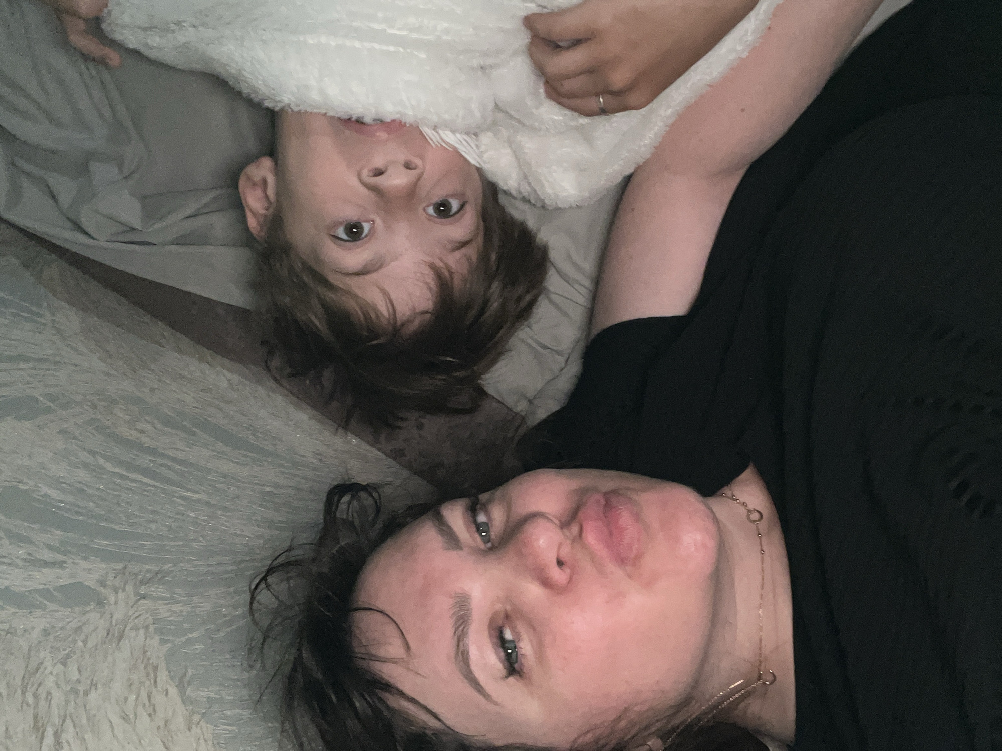 The image shows two individuals lying on a bed. One appears to be a child, possibly a young boy, partially covered by a white blanket. The child is looking directly at the camera. The second individual appears to be an adult woman. She is wearing a black shirt and a gold necklace. Her face is close to the camera and she is making a kissy face. The bed has patterned linens. The location information is not determinable from the image.