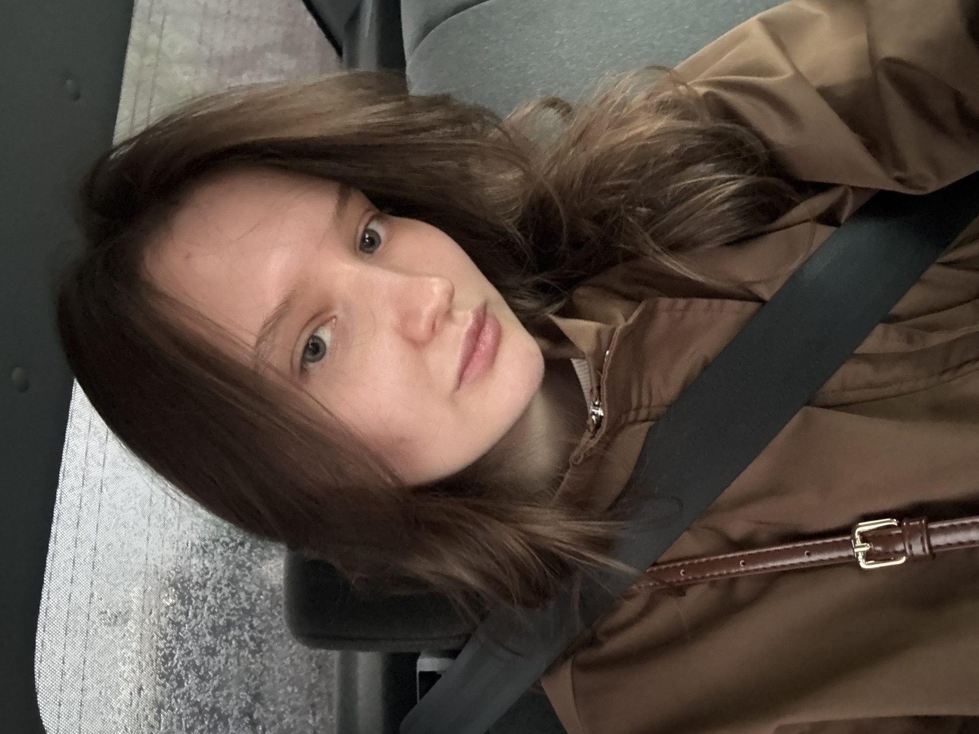 A young woman with light skin and brown hair is wearing a brown jacket and a seatbelt. The background shows a car window with raindrops, suggesting a rainy environment. The location is not discernible from the image provided, and cannot be confirmed as Khimki, Russia without additional information or context.