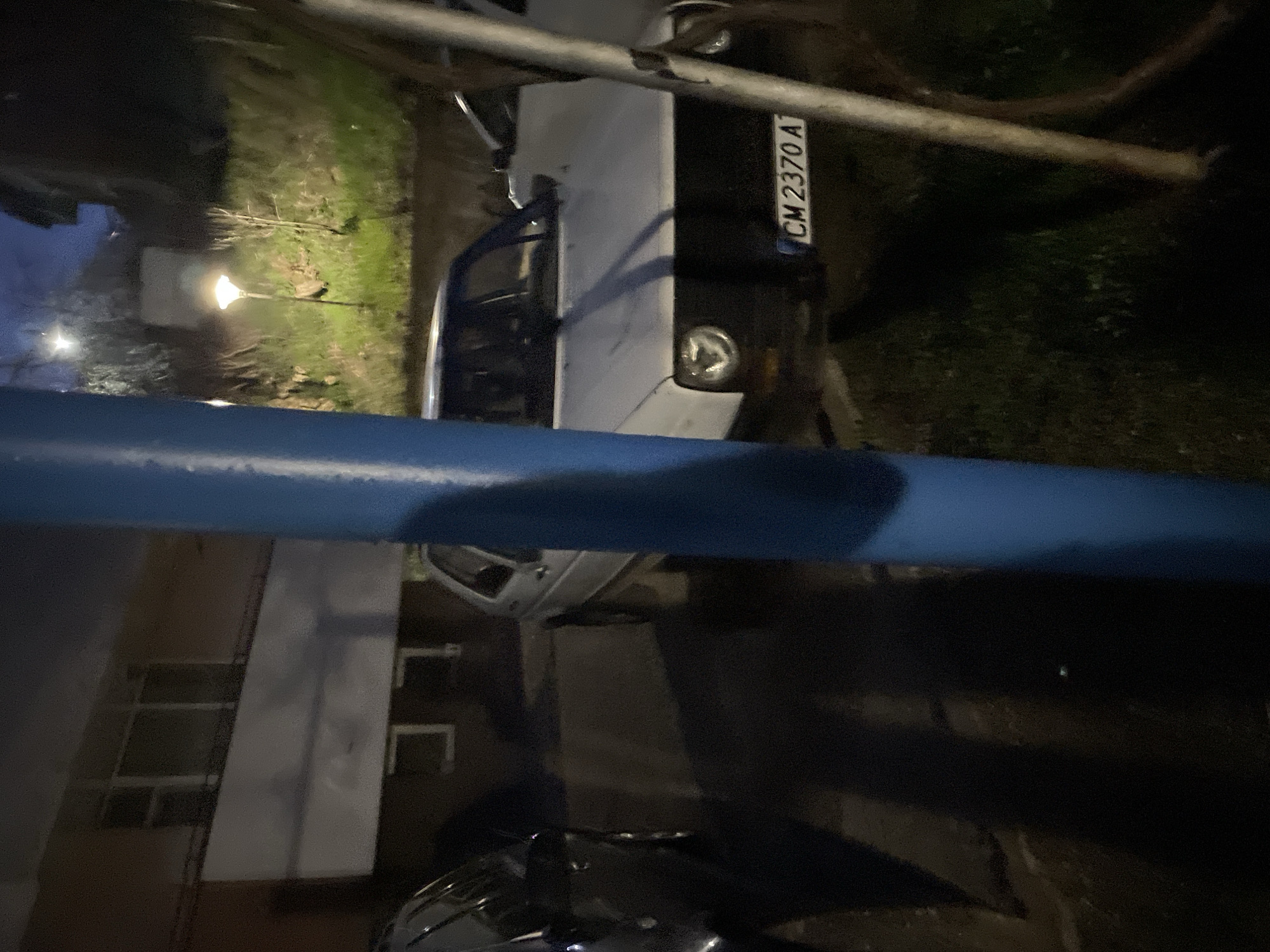 The image shows a street scene in Zlatograd, Bulgaria at night. A white Lada car with license plate "CM 2370 A" is parked on the right side of the frame. A streetlight is visible in the background. A blue pipe or railing is in the foreground, partially obscuring the view. A building with windows is visible on the left. A motorcycle is parked on the lower part of the image.