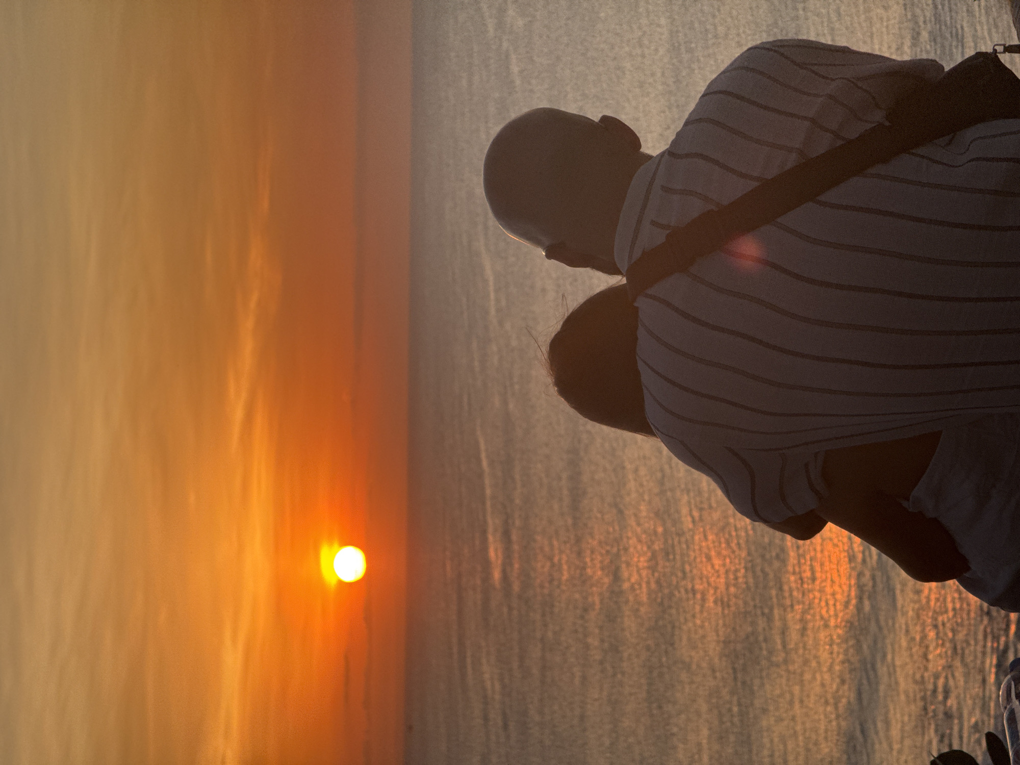 Here is a detailed description of the image:
The image shows two individuals silhouetted against a vibrant sunset. The figures, possibly a couple, are positioned on the right side of the frame, looking out towards the sea. One figure is bald, and both are wearing light-colored clothing with a striped pattern. A strap is visible across the shoulder of one individual.
The setting sun dominates the left side of the image, casting a warm, golden glow across the sky and water. The water reflects the colors of the sunset, creating a shimmering effect. The image composition suggests the subjects are observing this natural spectacle. The specific location is unknown.