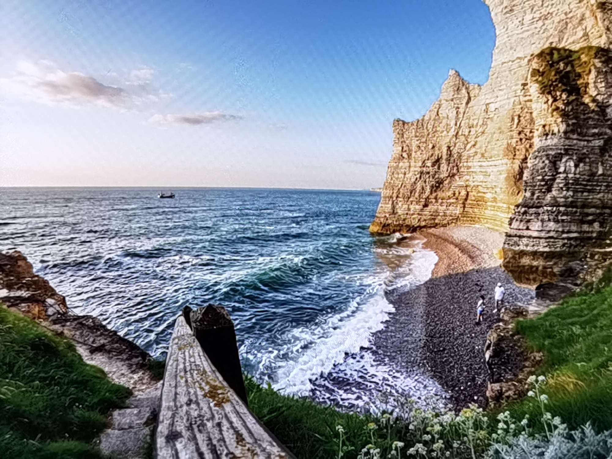 A wide-angle view captures a rugged coastline with towering chalk cliffs under a bright blue sky with a few wispy clouds. The ocean, a deep blue, is active with moderate waves crashing onto a pebble beach. The sun is likely high in the sky or mid-afternoon, casting shadows that highlight the textures of the cliffs and the white foam of the waves.
On the beach, two figures are visible, appearing small against the immense cliffs. They seem to be casually walking or standing, perhaps enjoying the scenery. A small boat is a distant speck on the water.
In the foreground, a wooden railing and stone steps suggest a path or viewpoint overlooking the beach and sea. Green grass and wildflowers grow on the cliff edges, adding a touch of life to the dramatic landscape. The overall scene evokes a sense of natural beauty, grandeur, and peaceful contemplation. The location is identified as Étretat, France, known for its distinctive rock formations. There is no visible text in the image.