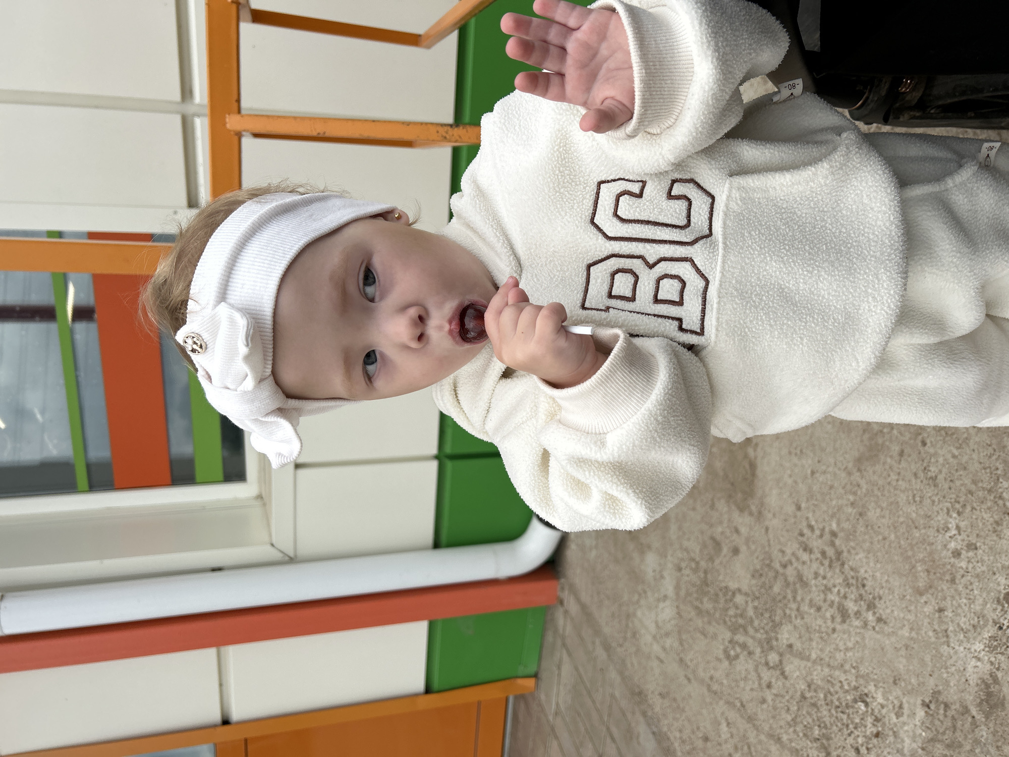 A young child with fair hair, wearing a white fuzzy hoodie and a white headband with a bow, is standing outside. The child is holding a dark-colored lollipop to their mouth and has their left hand raised, palm facing forward. The background shows a building with orange and green accents, as well as a grey, textured ground. The overall scene appears to be a candid photograph taken in a city environment, possibly near a playground or public space.