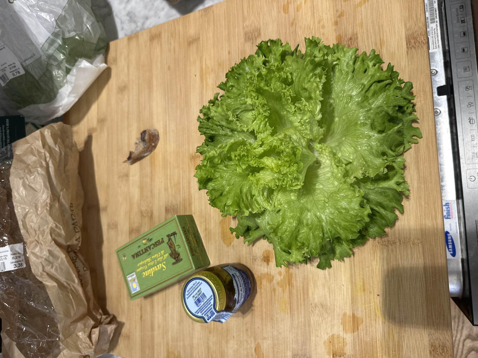 The image shows a wooden cutting board with various food items on it. A head of lettuce is positioned in the upper right quadrant. To the left and slightly below the lettuce is a green box of "Sardine" by "Pescantina" next to a small jar of "Filetti di Acciughe" (anchovy fillets). There is a partially visible piece of what appears to be bread or a crouton on the left side of the lettuce. In the lower left corner, there is a torn brown paper bag with part of a plastic bag. In the upper left, there is a partially visible bag of lettuce.
The edge of a Samsung dishwasher is visible on the right side of the image. Labels on the packaging indicate Italian text, specifically mentioning "Olio Extra Vergine," "Prodotto in," and "Cliva Biologico" and labels with "aturaslit" and price markings. The brand “Finish” is visible on the dishwasher.
It's impossible to determine the location in the city of Milan, Italy, from the image alone. The scene is set on a cutting board, likely in a kitchen.