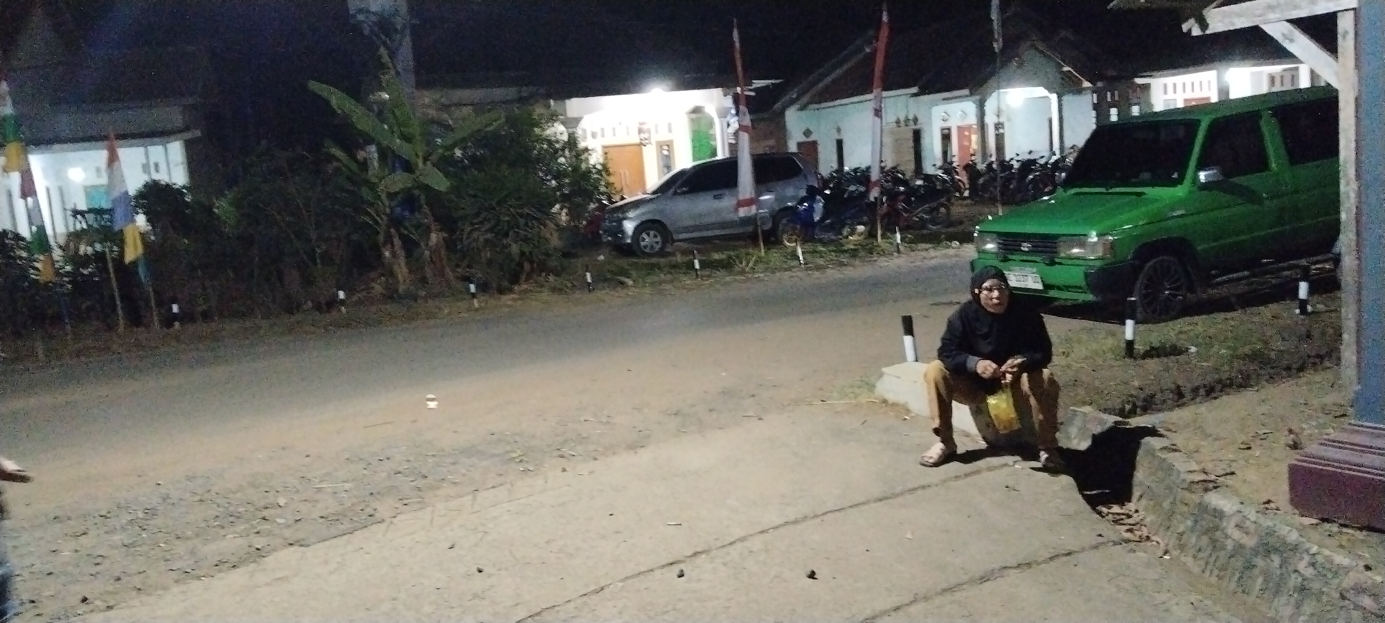 In the city of Sido Rahayu, Indonesia, a person wearing a black headscarf and black jacket sits on the side of a paved road at night. The individual is also wearing light brown pants and sandals, and is holding a yellow bag in their lap. Behind the person, a bright green van is parked on the side of the road, and several motorcycles are visible further back. The scene appears to be in a residential area, with houses and foliage bordering the road. The lighting suggests it is nighttime, with artificial lights illuminating the area.