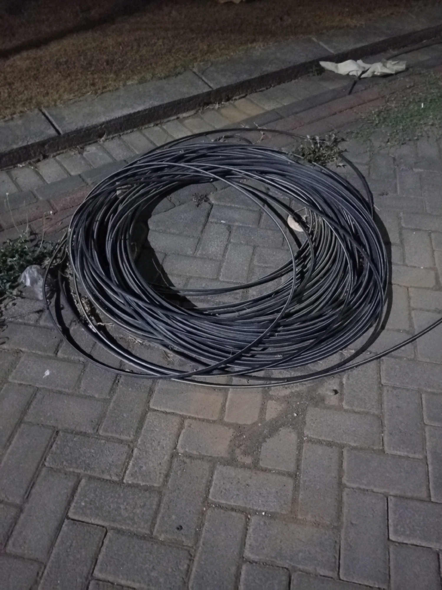 A coiled black cable lies on a paved area, likely a sidewalk or pathway, made of interlocking brick pavers. The ground is lit by artificial light, casting shadows. To the left of the cable, there is a strip of dry grass or ground cover bordered by a concrete edge. In the background, a patch of dry grass is visible, with some scattered debris. A white plastic bag is partially visible near the grass. The scene suggests an outdoor urban setting in Malang, Indonesia, possibly during the night or evening due to the lighting conditions.