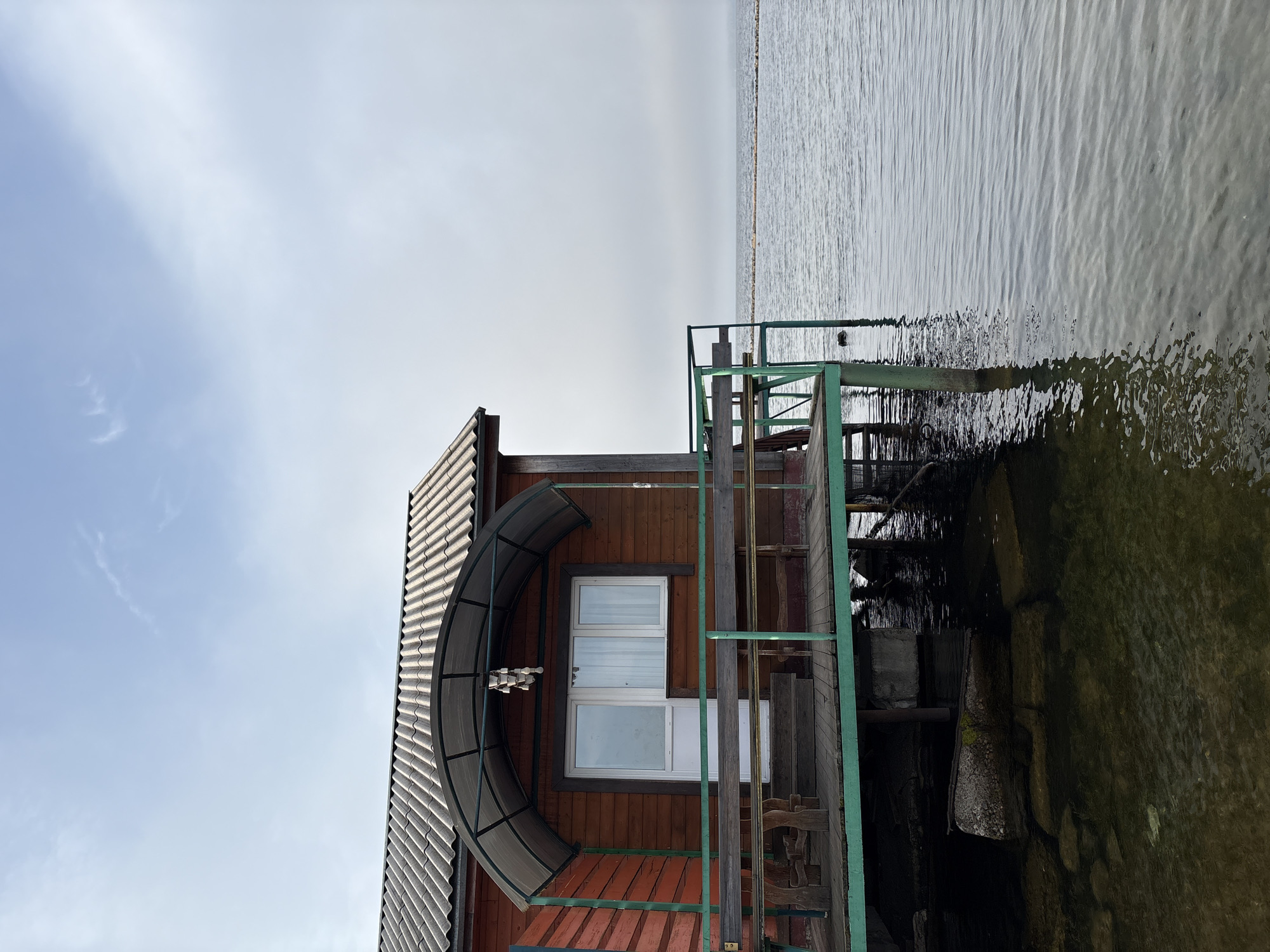 A wooden, red-colored building stands on stilts over the water. The building has a gray, corrugated roof and a covered porch with a translucent roof. A white door and a window are visible on the front of the building. A wooden deck with benches and a table extends from the front of the building. The water appears calm, with some rocks visible beneath the surface near the building. In the distance, a thin line of land or a breakwater is visible on the horizon. The sky is overcast with a few wispy clouds. The scene is located in Ust-Ilimsk, Russia.