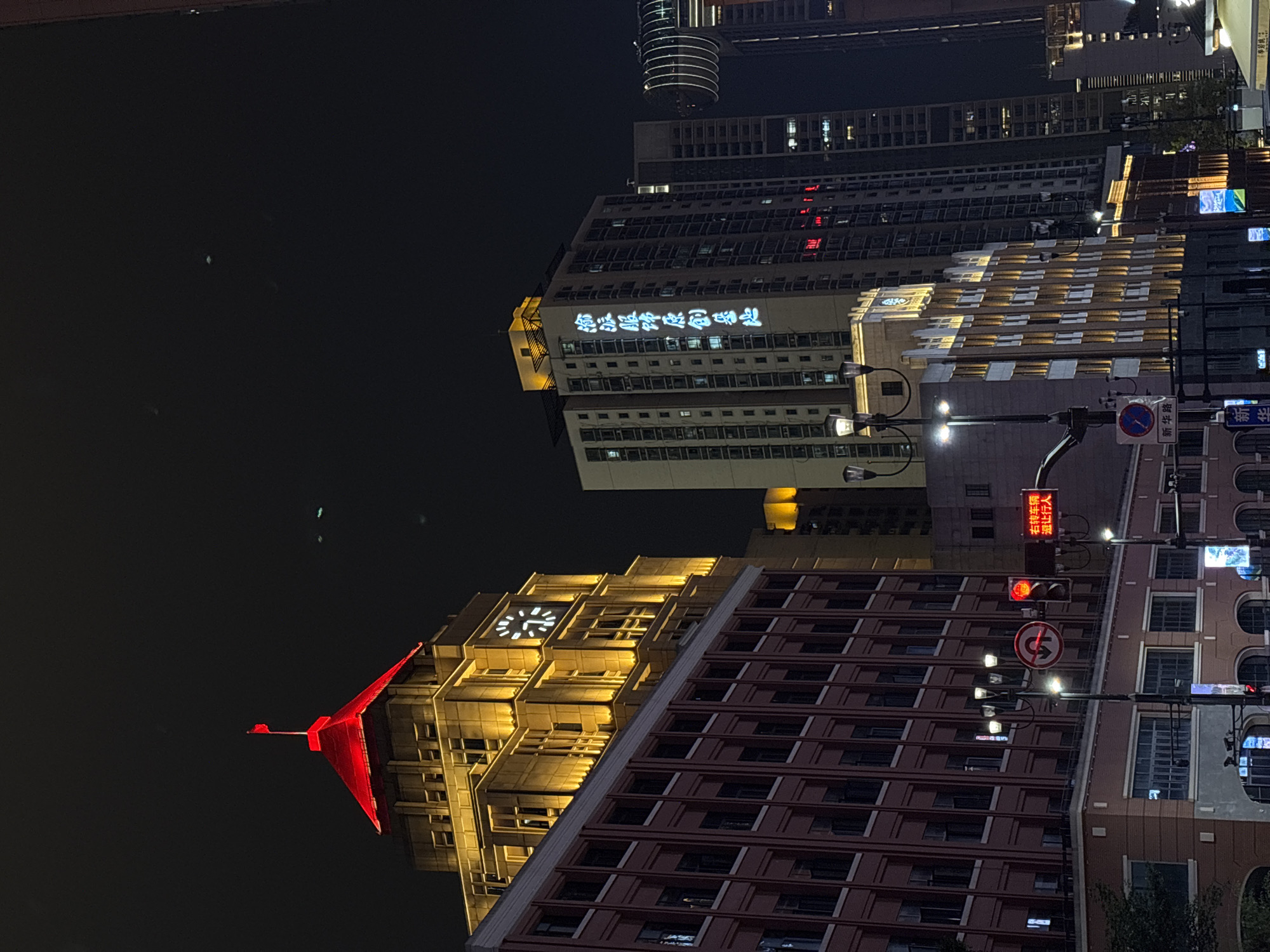 The image depicts a vibrant urban cityscape at night in what appears to be Yuzhong District, China. The scene is dominated by several tall buildings, brightly illuminated against a dark, clear sky.
In the left foreground, a prominent feature is a large, striking red sculpture, resembling a bird with outstretched wings, perched atop a building that also displays an illuminated clock face. The clock's hands suggest a time close to midnight or 1 AM.
A central, multi-story building features bright blue neon Chinese characters spelling out "新华酒店" (Xinhua Hotel). Other buildings in the background and to the right display various architectural styles, with some facades lit by warm yellow uplighting and others showing numerous lit windows.
The lower and middle sections of the image include urban infrastructure such as streetlights, which cast bright halos, and traffic lights. Visible traffic signs provide additional context: a blue and white sign clearly reads "新华路" (Xinhua Road). Above a traffic light, a red illuminated sign displays Chinese characters that translate to "右转车辆 温让行人" (Right-turning vehicles, yield to pedestrians). Additionally, a circular white sign with a red border and a black arrow indicates a "No U-turn" restriction. Several traffic lights are illuminated red.
The overall impression is of a bustling city center after dark, characterized by a dynamic blend of modern architecture, prominent signage, and active street infrastructure, despite no people being explicitly visible.