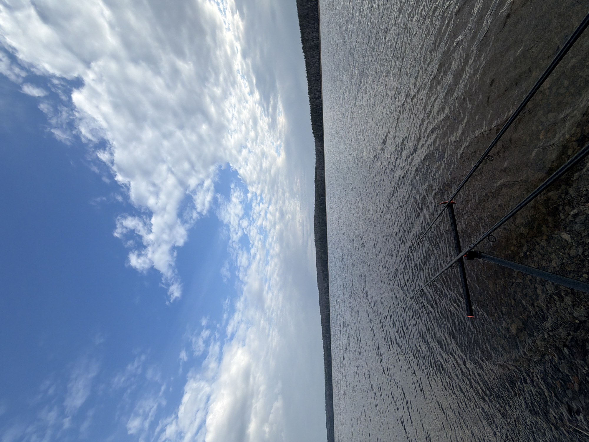The image shows a body of water, possibly a lake or river, with fishing rods in the foreground. The shoreline consists of rocky ground. A line of trees or a forest appears on the horizon separating the water from the sky. The sky is divided into areas of blue with scattered clouds and areas of dense, gray clouds. The location is possibly in the city of Chaikovsky, Russia. There are no people visible. The scene is calm and serene.