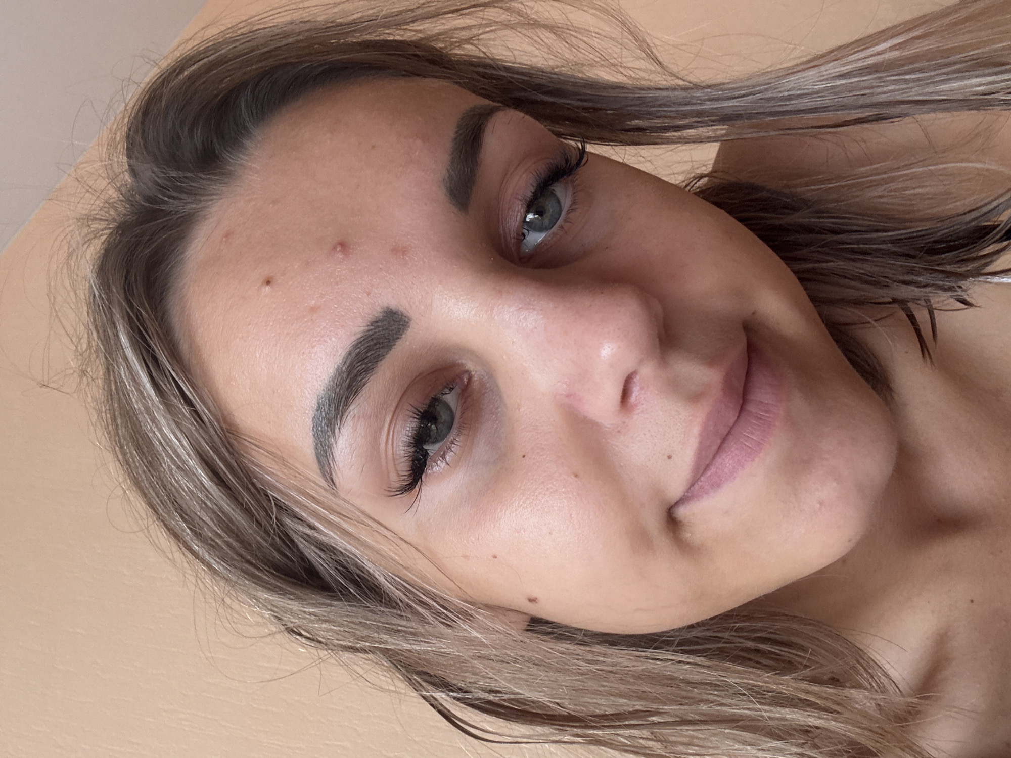 A close-up, head-and-shoulders selfie of a young woman with light brown hair and blue eyes. She is looking towards the camera with a slight smile. The background is a plain, light orange wall. The woman has light makeup on, including mascara, eyeliner, and lipstick. There are a few blemishes visible on her forehead. Her hair is slightly wet and styled loosely around her face. The lighting is natural and appears to be from an overhead source. There are no identifiable landmarks or contextual clues within the image that would suggest a specific location in Kholmsk, Russia, or any other city. The image is a personal portrait.