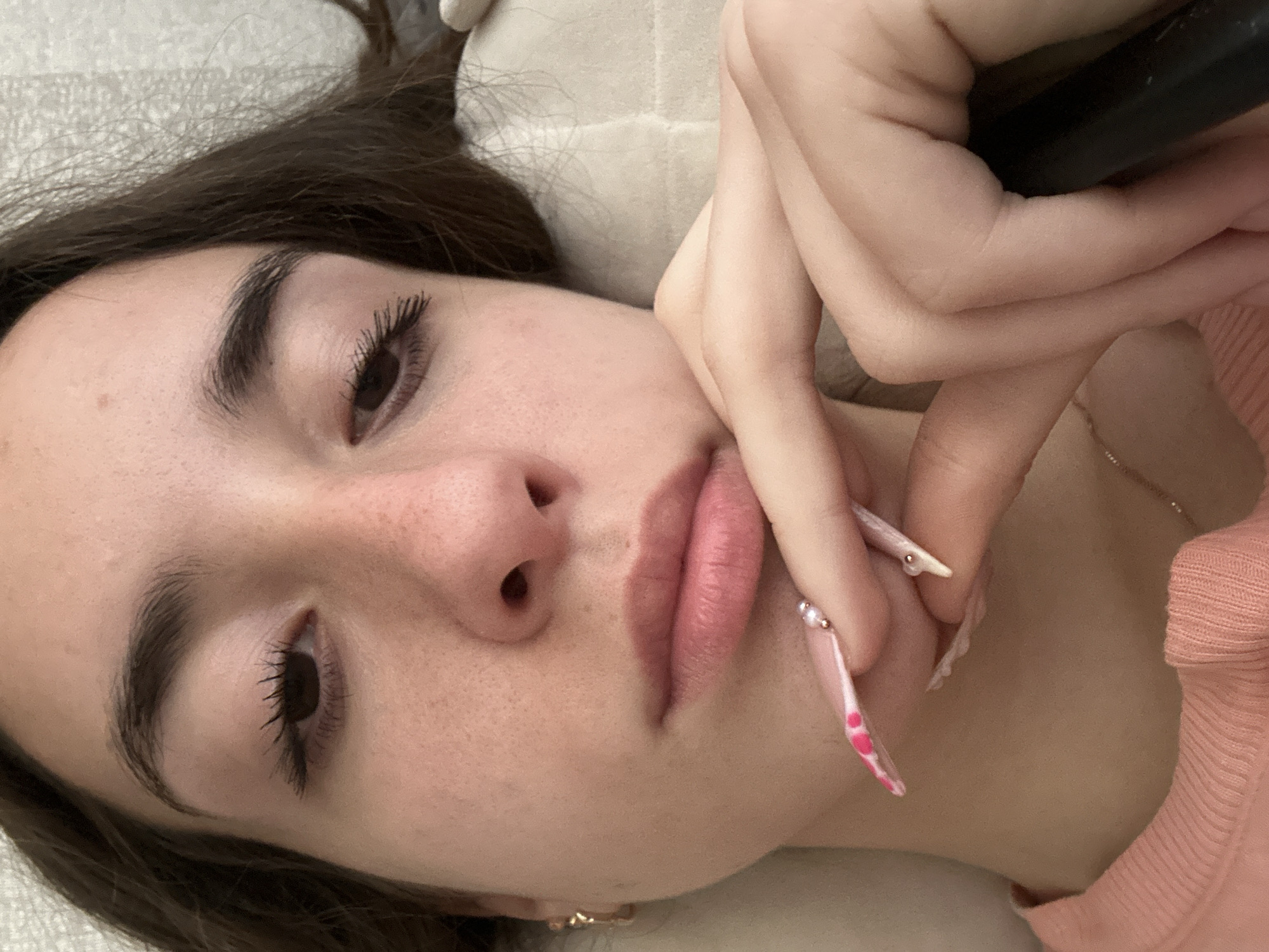 A close-up, slightly angled portrait of a young woman with dark hair, fair skin, and freckles is captured in an indoor setting. Her right eye is visible, adorned with dark mascara. Her lips are slightly parted, and her chin rests on her right hand, which features long, pink-tipped acrylic nails with small pearl embellishments. She is wearing a light pink, ribbed long-sleeved shirt. The background appears to be a textured, light-colored surface, possibly a pillow or bedding. The image does not provide any specific details or context related to the city of Shumen, Bulgaria.