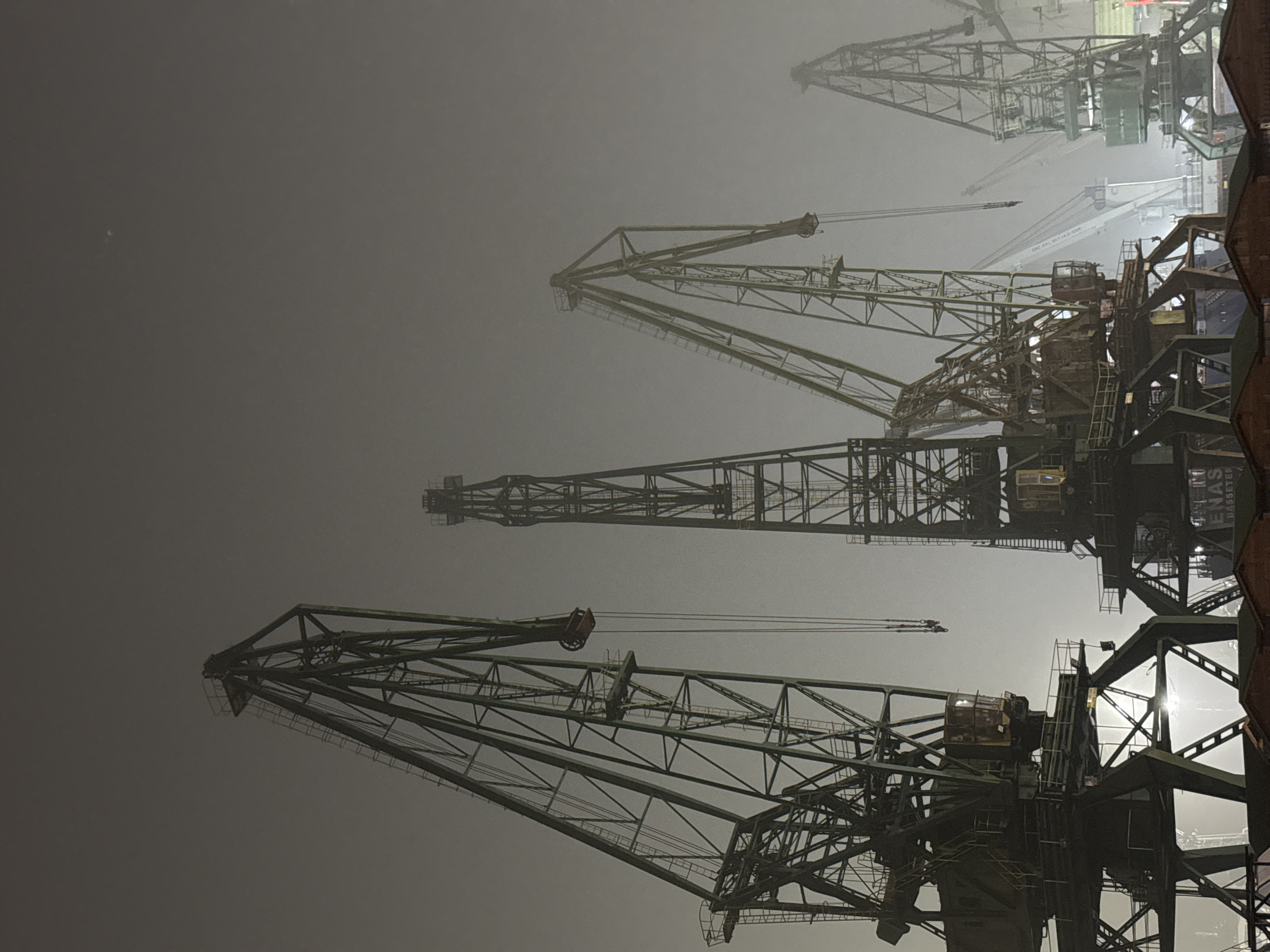 The image shows several large industrial cranes at the Port of Varna, Bulgaria, shrouded in heavy fog or mist. The cranes appear to be of older design, with a lattice structure. Some are closer to the viewer and appear larger, while others recede into the fog, suggesting depth. The illumination is uneven, with some cranes showing brighter highlights where light penetrates the fog and others being in deep shadow. The overall atmosphere is muted and indistinct due to the fog.