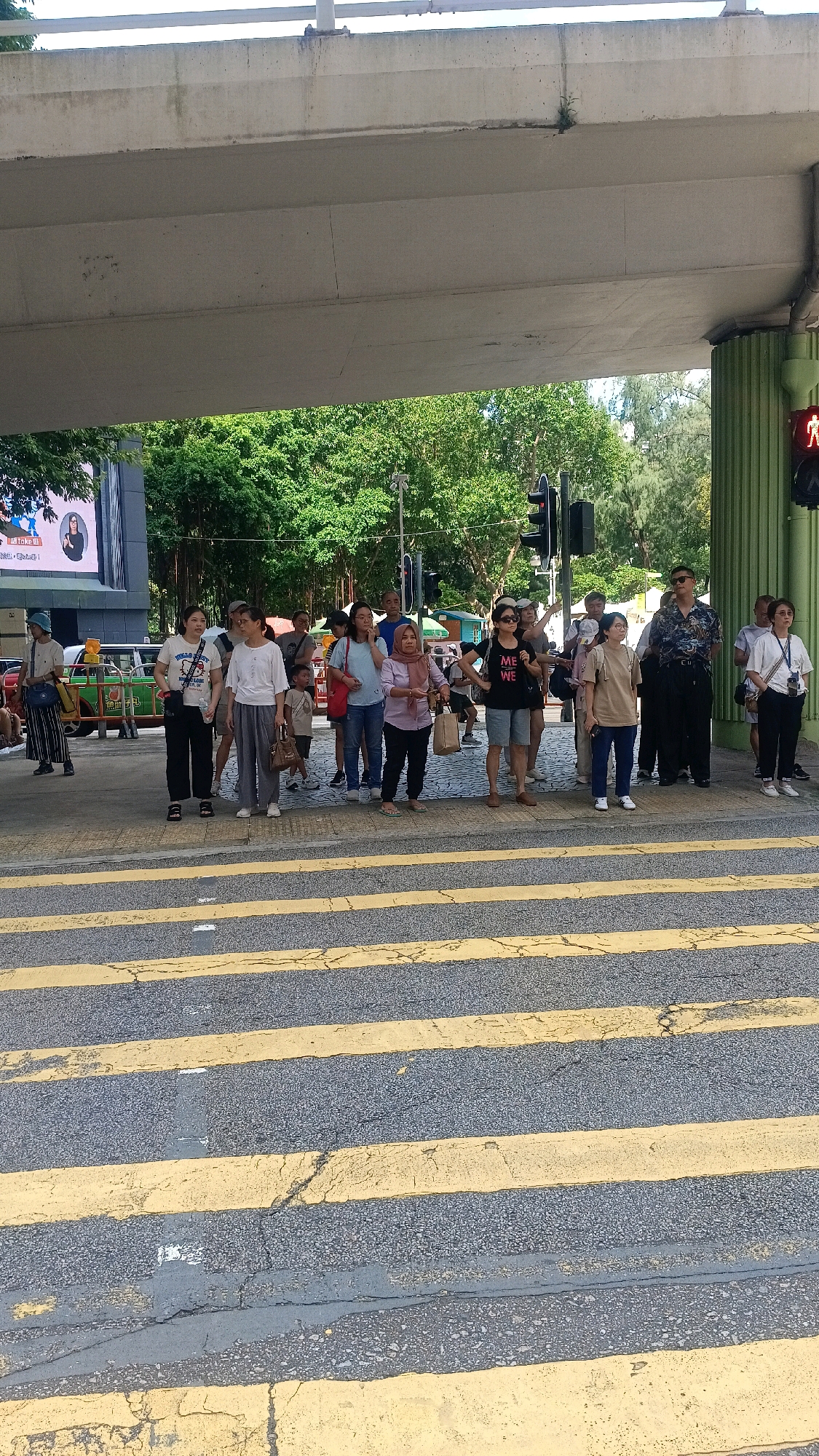 The image shows a group of people waiting at a pedestrian crossing under an elevated road structure. The pedestrian crossing is marked with yellow and grey stripes on the asphalt. In the background, there are trees and a green building with some scaffolding. A traffic light is visible, showing a red signal for pedestrians, indicating a "don't walk" symbol. Several individuals, including men, women, and at least one child, are standing on the sidewalk, some looking towards the street, others engaged in conversation. One person in the foreground is wearing a black t-shirt with the text "ME WE" printed on it. The overall scene suggests an urban environment, likely in Hong Kong, China, given the architecture and the presence of a pedestrian crossing with specific traffic signals.