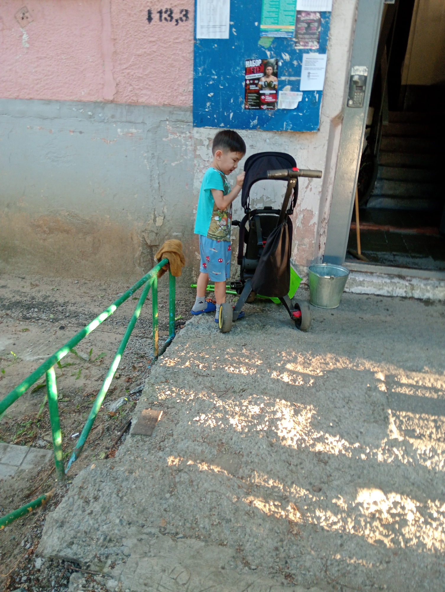 In an outdoor urban setting, a young boy stands beside a black stroller. He is wearing a turquoise t-shirt and blue shorts with a cartoon pattern. The stroller has a silver handle and a black fabric seat. To the left of the boy, a green metal railing extends into the foreground. Behind the stroller, a silver bucket rests on the ground. The background features a textured, pale pink wall with a grey concrete base. A blue bulletin board is affixed to the wall, displaying various posters. To the right of the stroller, a dark entryway with stairs is visible. The ground is a mix of concrete and gravel, with shadows cast by sunlight. The image appears to have been taken during the daytime.