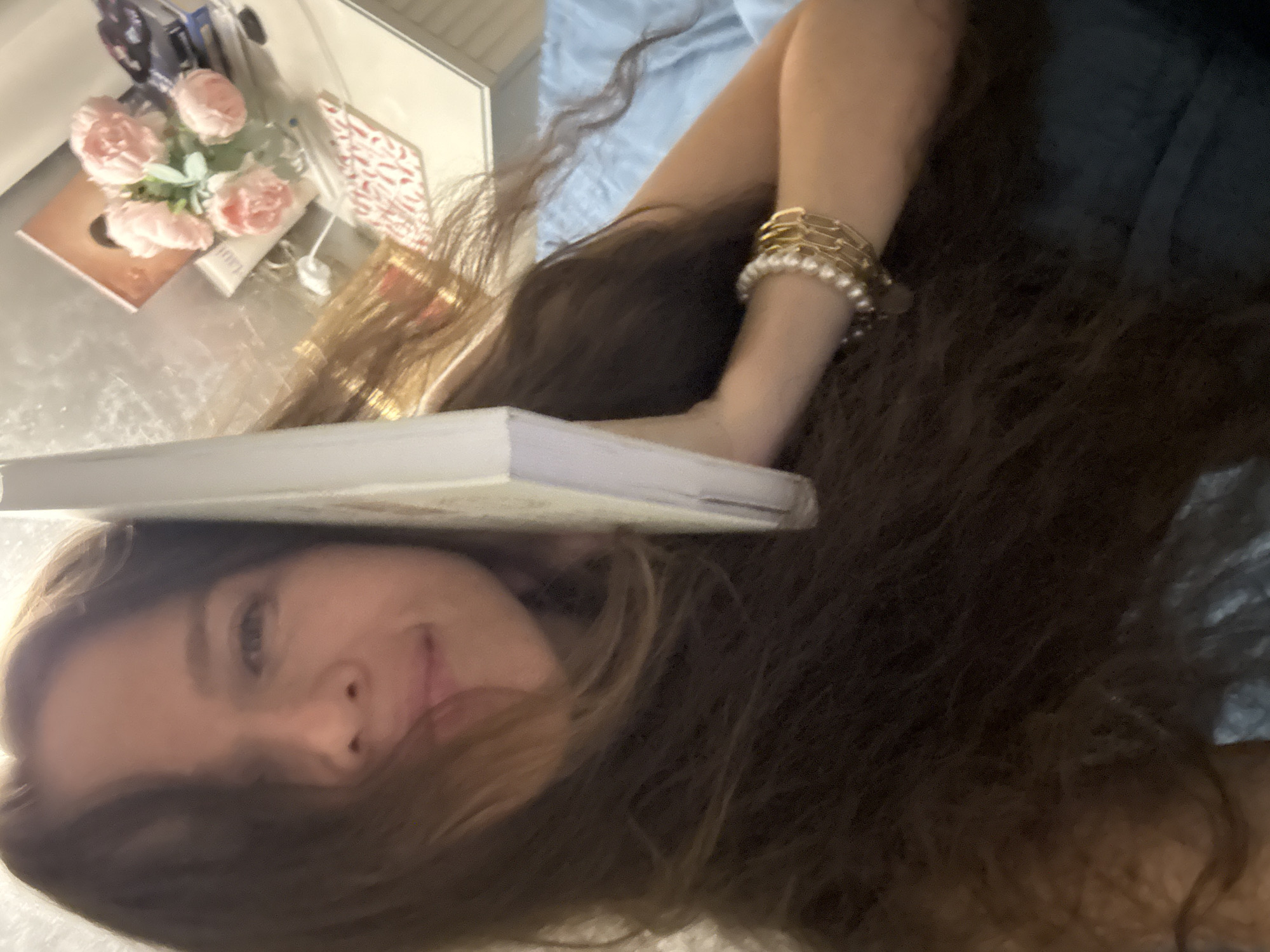 A woman with long brown hair is lying down, holding a white book to the side of her face. She is wearing a pearl and gold bracelet on her left wrist. The background shows a bedside table with decorative pink flowers, a book, and other small items. The overall scene appears to be indoors, likely in a bedroom. No specific location in Khimki, Russia is discernible from the image.