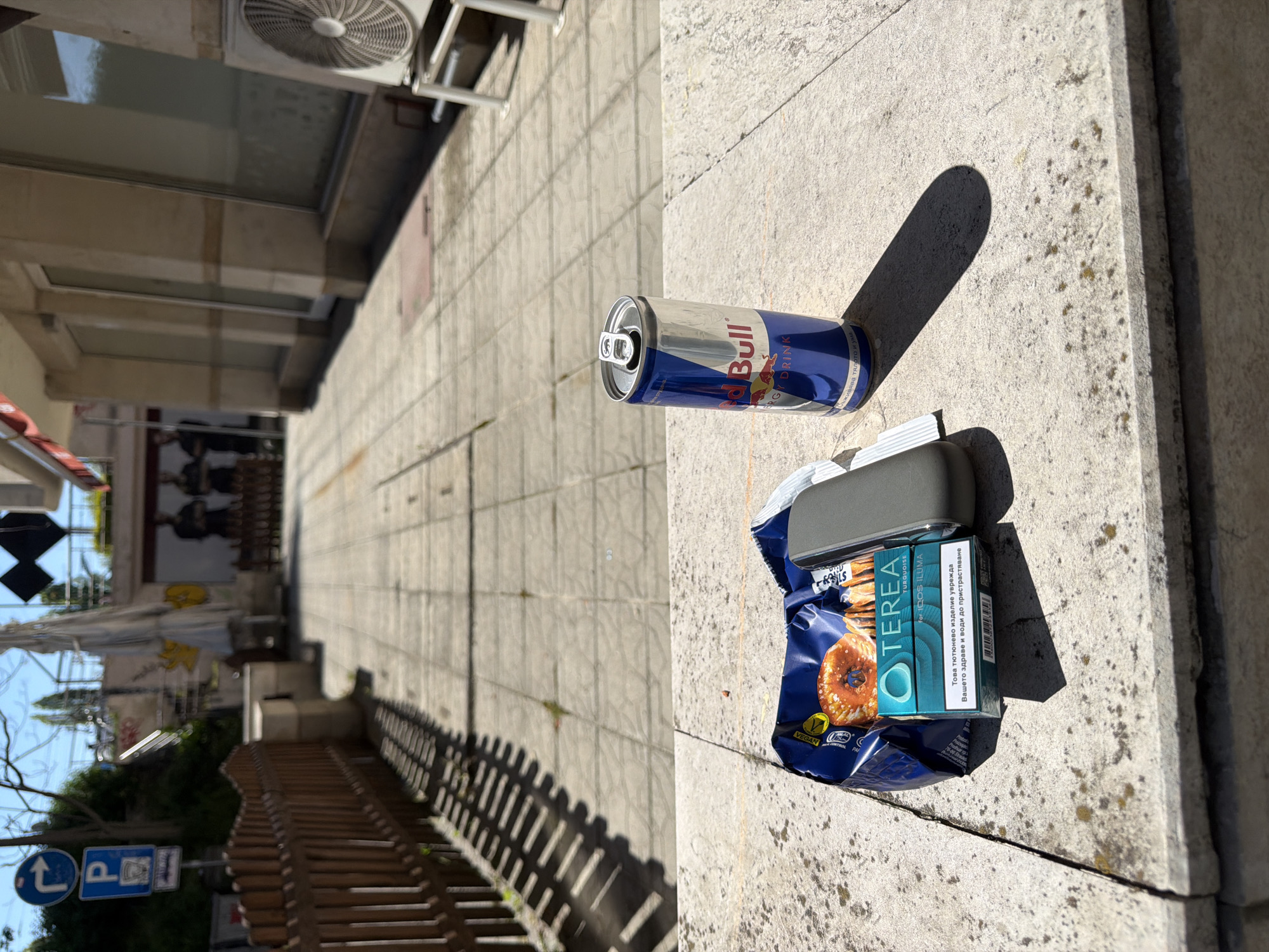 The image depicts litter on a concrete sidewalk in Sofia, Bulgaria. Litter objects: a partially-consumed can of Red Bull energy drink, a blue snack bag (labeled "Vegan Rolls" and bearing an image of a donut), a closed pack of TEREA Turquoise heat sticks for IQOS ILUMA.
The background shows a building with glass windows and awnings, part of an air conditioning unit. Street signs are visible further down the street, partially obscured by trees and a fence.