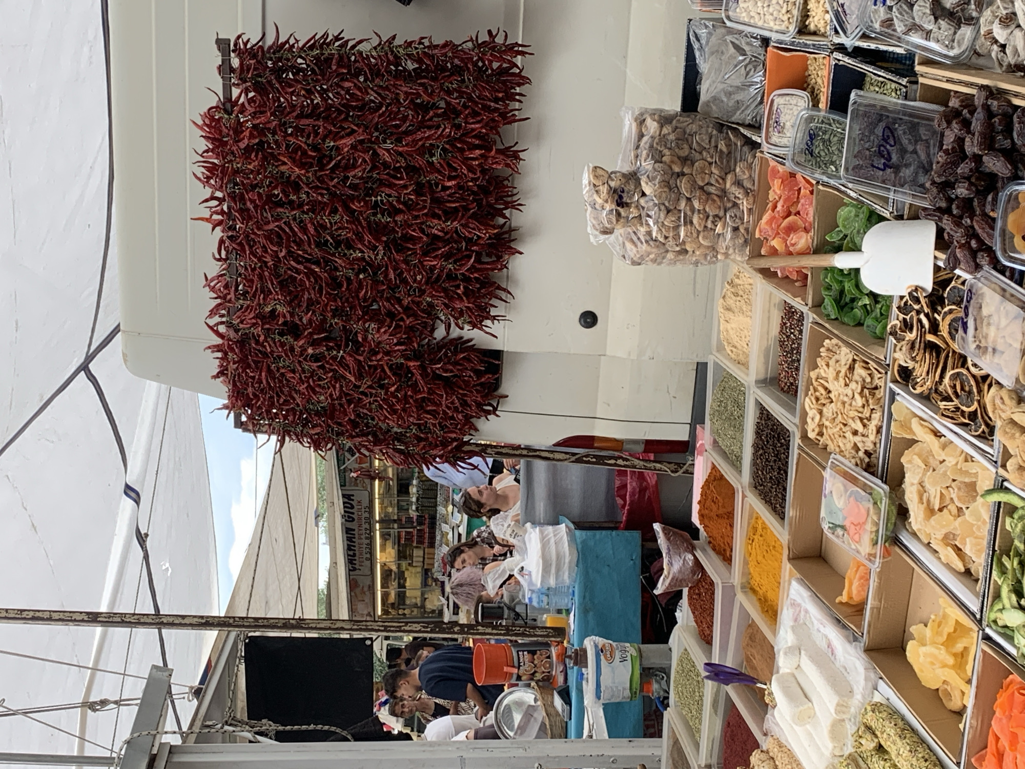 The image shows a market stall in Fethiye, Turkey, displaying a variety of goods. On the left, a large collection of dried red peppers hangs from the stall's structure. To the right of the peppers, various dried fruits, nuts, and spices are organized in individual containers or bins. Customers and vendors are visible in the background. The stall is partially covered by a white canopy or awning.