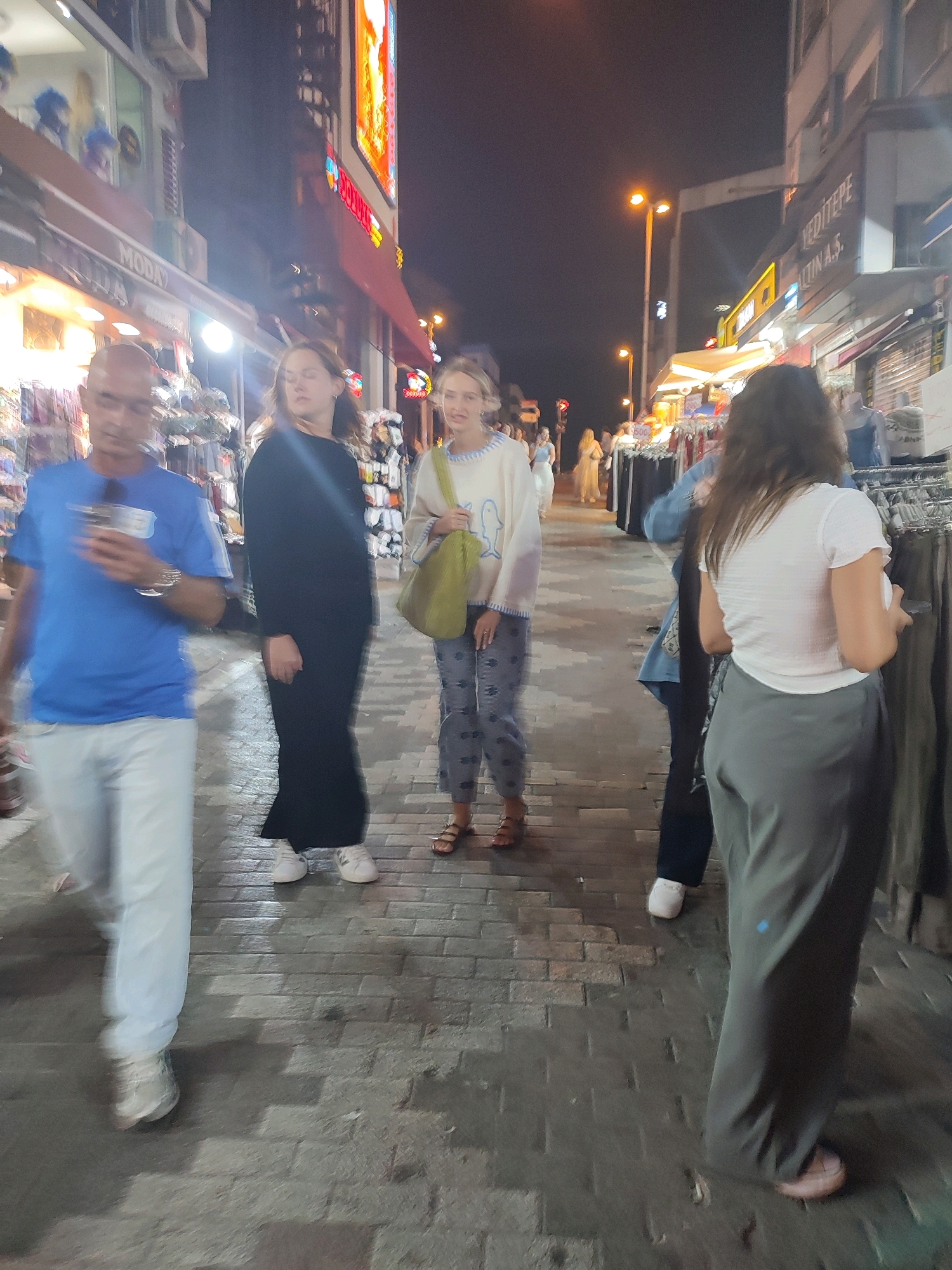 The image depicts a street scene at night in Kadıköy, Turkey, characterized by the presence of shops and pedestrian activity. The foreground features a man wearing a blue t-shirt and white pants, walking with a phone in his hand. To his right is a woman in a long black dress and white sneakers, looking forward. Further to the right, a woman in a light-colored sweater and patterned pants is seen holding a green bag. The background shows a street lined with shops, some with illuminated signs, including one with the text "MODA". Streetlights and overhead lighting illuminate the scene, casting shadows on the brick-paved walkway. The overall atmosphere suggests a lively commercial area at night.