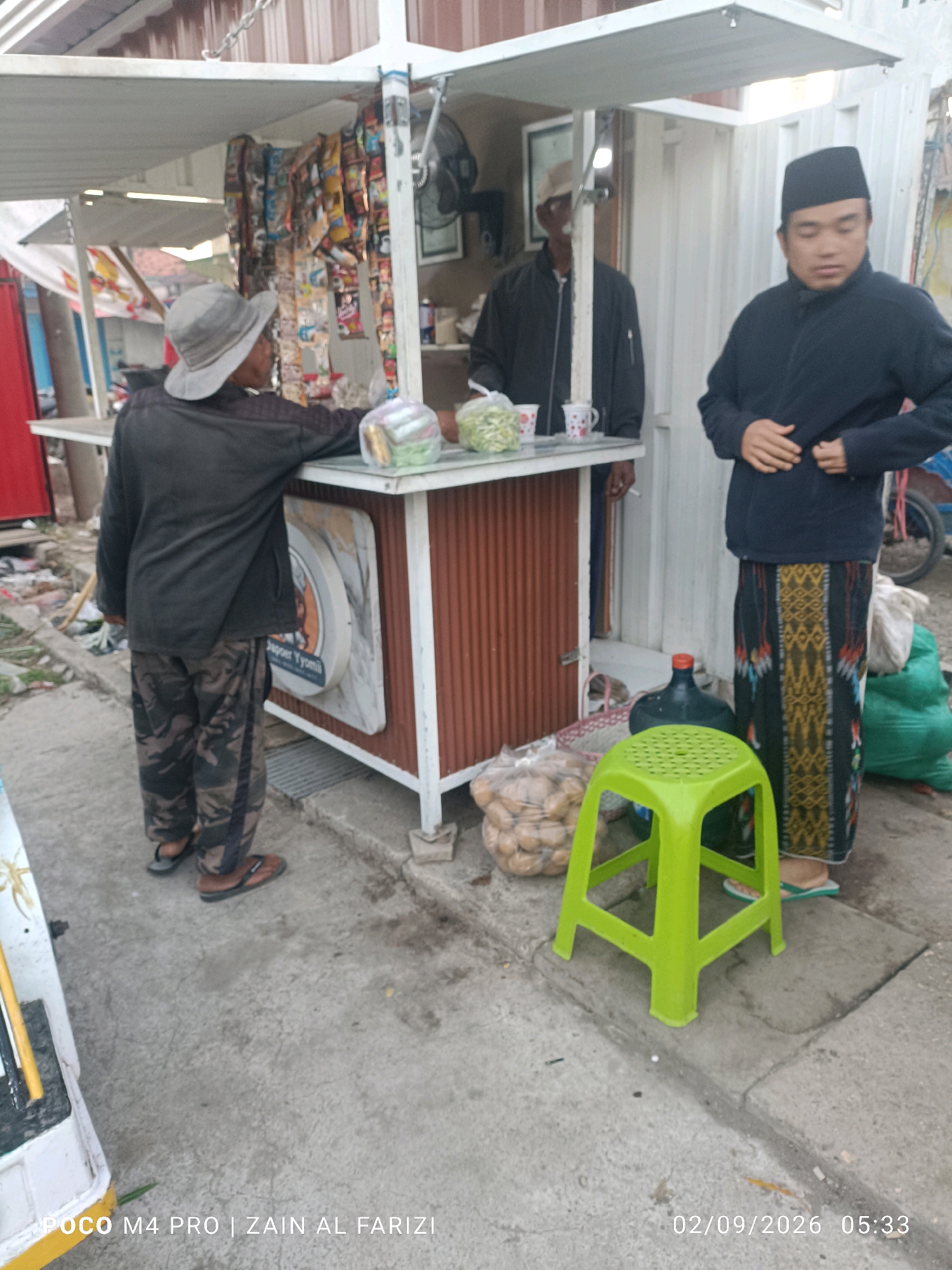 In the city of Arosbaya, Indonesia, a man wearing a grey hat and camouflage pants stands facing a food stall. He leans on the counter, looking at items displayed on it. A second man in a black jacket and a peci (a cap commonly worn by Muslim men in Indonesia) stands to the right of the stall, adjusting his clothing. Behind them, a third person, mostly obscured, appears to be working at the stall. The stall is stocked with packaged snacks and other goods. In front of the stall, a green plastic stool, a large water jug, and bags of food are visible on the ground. The scene appears to be an outdoor market or roadside vendor setting.