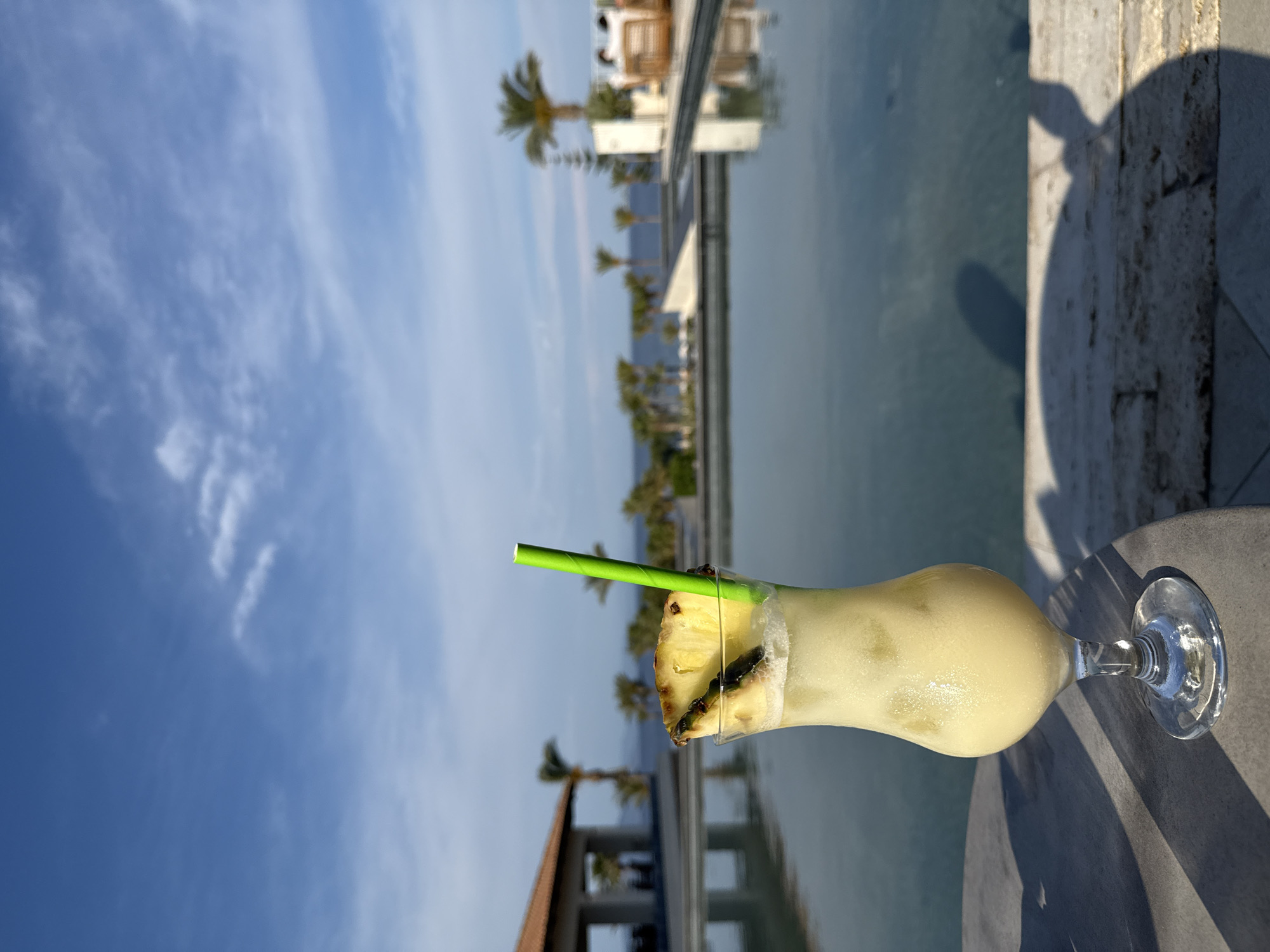 A medium shot captures a cocktail glass filled with a creamy yellow beverage, garnished with a slice of pineapple and a green straw. The glass rests on a round, gray table, casting a shadow. The background features a serene body of water reflecting the sky and surrounding architecture, including palm trees. The scene is set in the city of Kos, Greece.