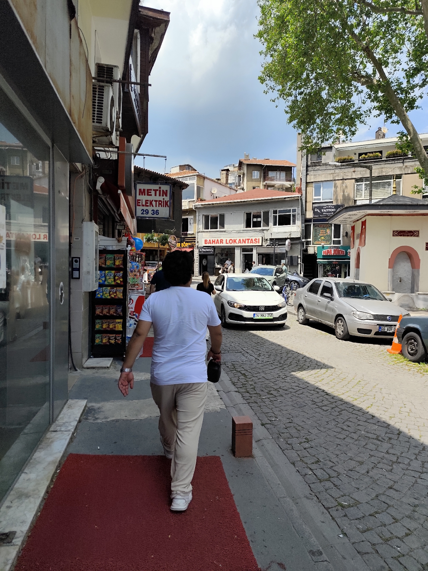 The image shows a street scene in Sarıyer, Turkey. A person wearing a white t-shirt and khaki pants is walking on the sidewalk to the right. A red mat is on the sidewalk in front of a shop. The street is paved with cobblestones, and several parked cars are visible. A sign that reads "BAHAR LOKANTASI" can be seen across the street. Another sign reads "METİN ELEKTRİK 29 36." A white building with a red-tiled roof is visible further down the street. Above that building, one can see a sign for "ISTINYE TATTOO STUDIO." A small stone structure resembling a fountain or well is to the right of the frame.