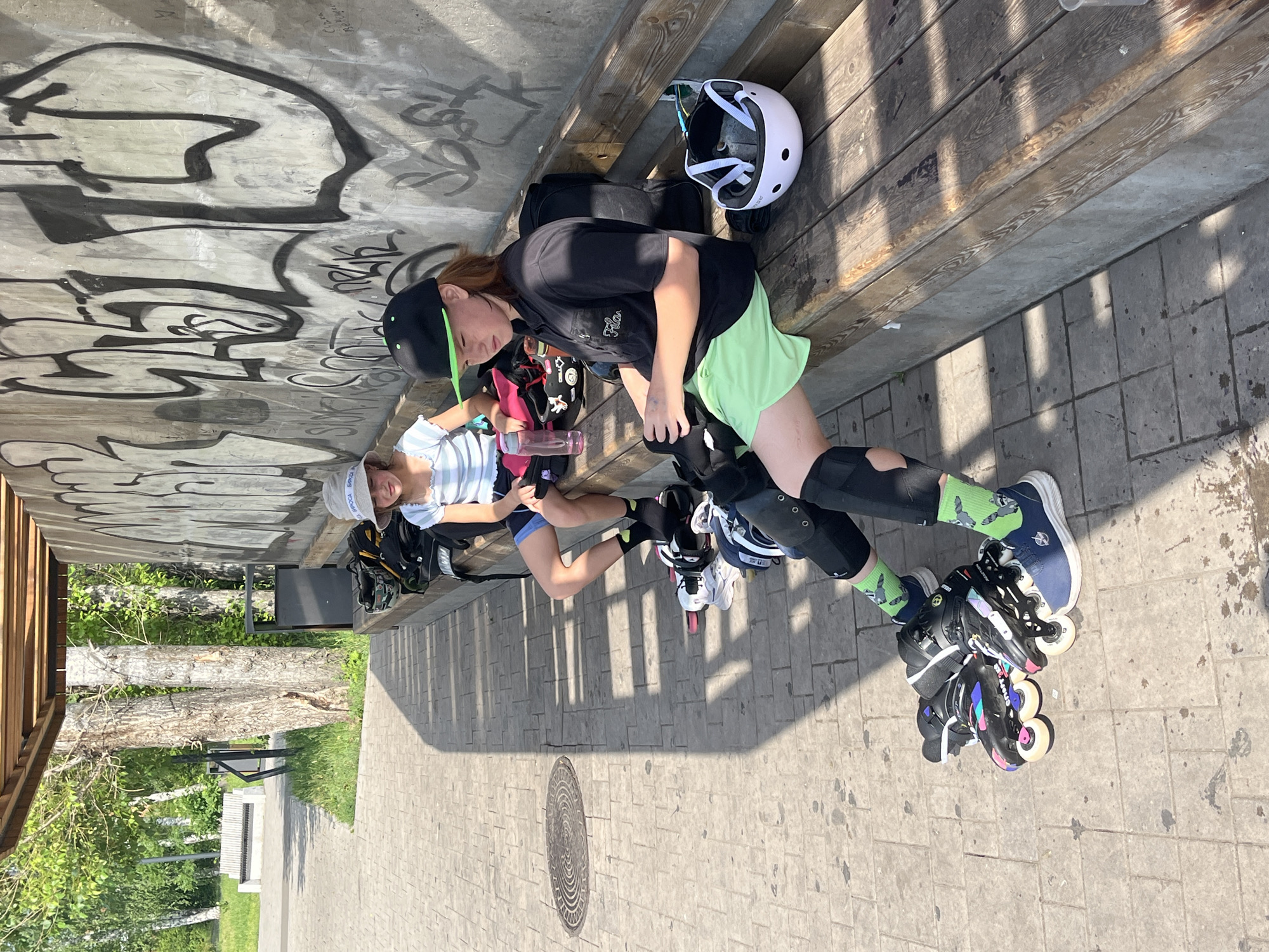 Two young individuals are seated on a wooden bench under a covered area with a graffiti-covered concrete wall behind them. The bench is in a park-like setting, indicated by trees and paved walkways. The individual on the left is wearing a light blue and white striped shirt, blue shorts, and a light-colored bucket hat. They are looking down and appear to be resting. The individual on the right is wearing a dark polo shirt, bright green shorts, a black baseball cap with a neon green brim, and black socks with a raccoon pattern. They have on inline skating protective gear including knee pads. Their inline skates are on the ground in front of them, along with another pair. A white and black helmet rests on the bench to their right. A pink water bottle is visible on the bench to the left of the first individual. The scene appears to be outdoors on a sunny day, with strong shadows cast on the ground. The location is identified as Divnogorsk, Russia.