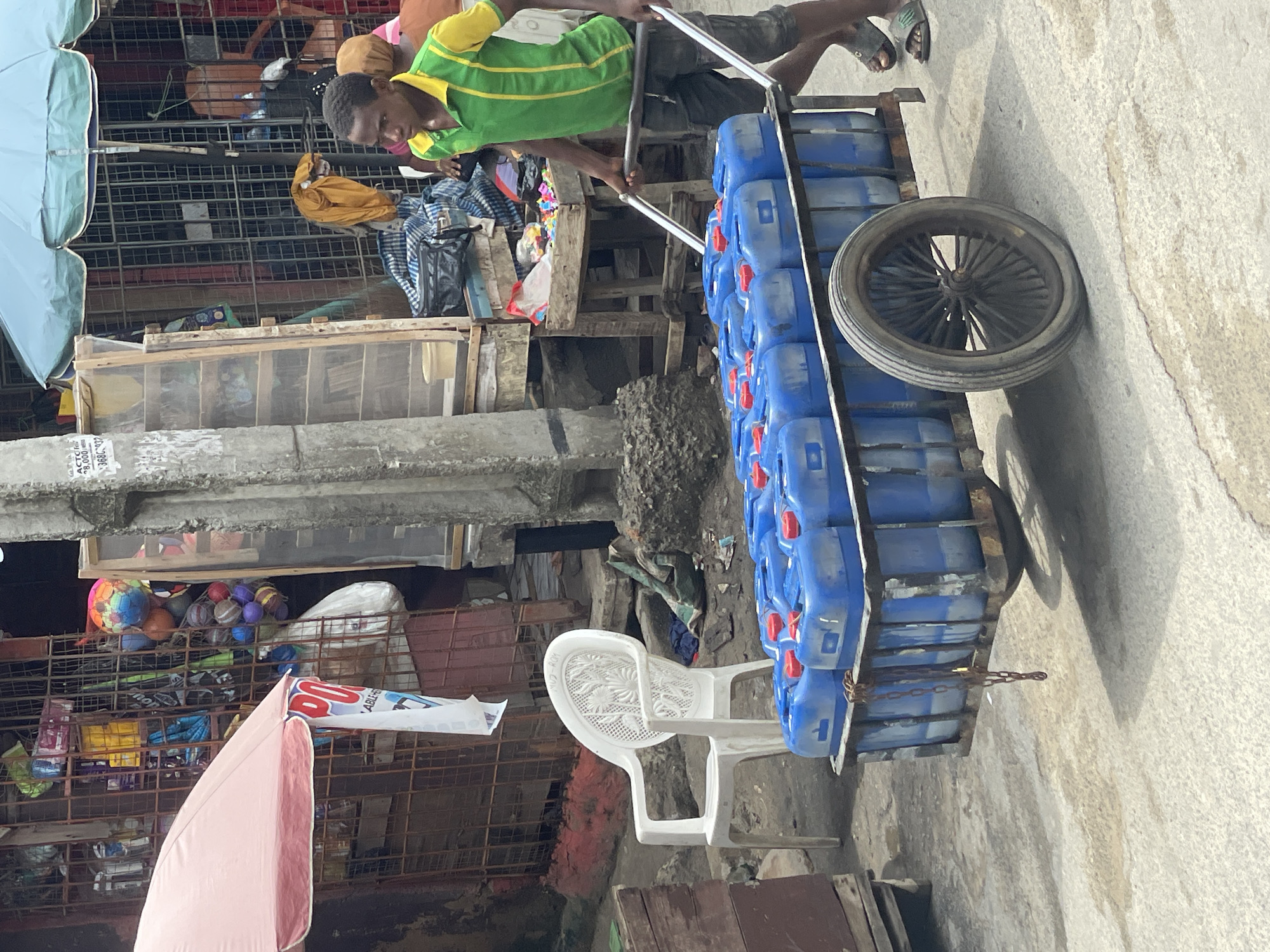 A man in a green and yellow shirt is pulling a cart loaded with blue jerrycans. The cart has a single large tire. The scene appears to be set in Lagos, Nigeria, indicated by the style of architecture, presence of water containers, the market scene in the background, and the overall atmosphere. The environment seems to be a bustling market or street, with various stalls and structures visible, a white chair, and an umbrella.