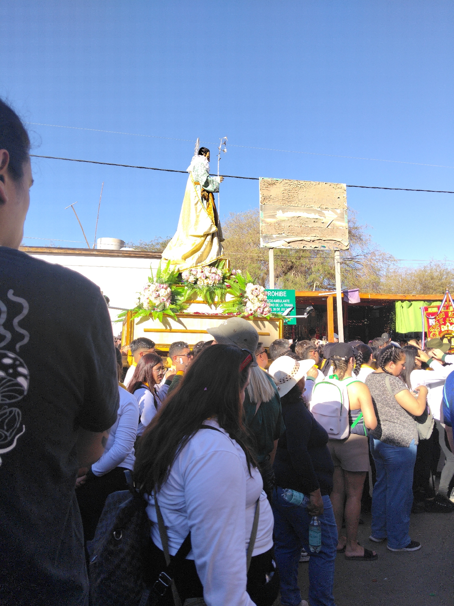In Pozo Almonte, Chile, a religious procession is underway, with a large statue of a saint, draped in gold fabric and adorned with flowers, being carried. A crowd of people surrounds the statue, some observing, others participating in the event. The sky is clear and blue, suggesting it is a sunny day. Buildings and trees are visible in the background, indicating an urban or semi-urban setting. The scene appears festive, with a sense of community and shared religious observance.