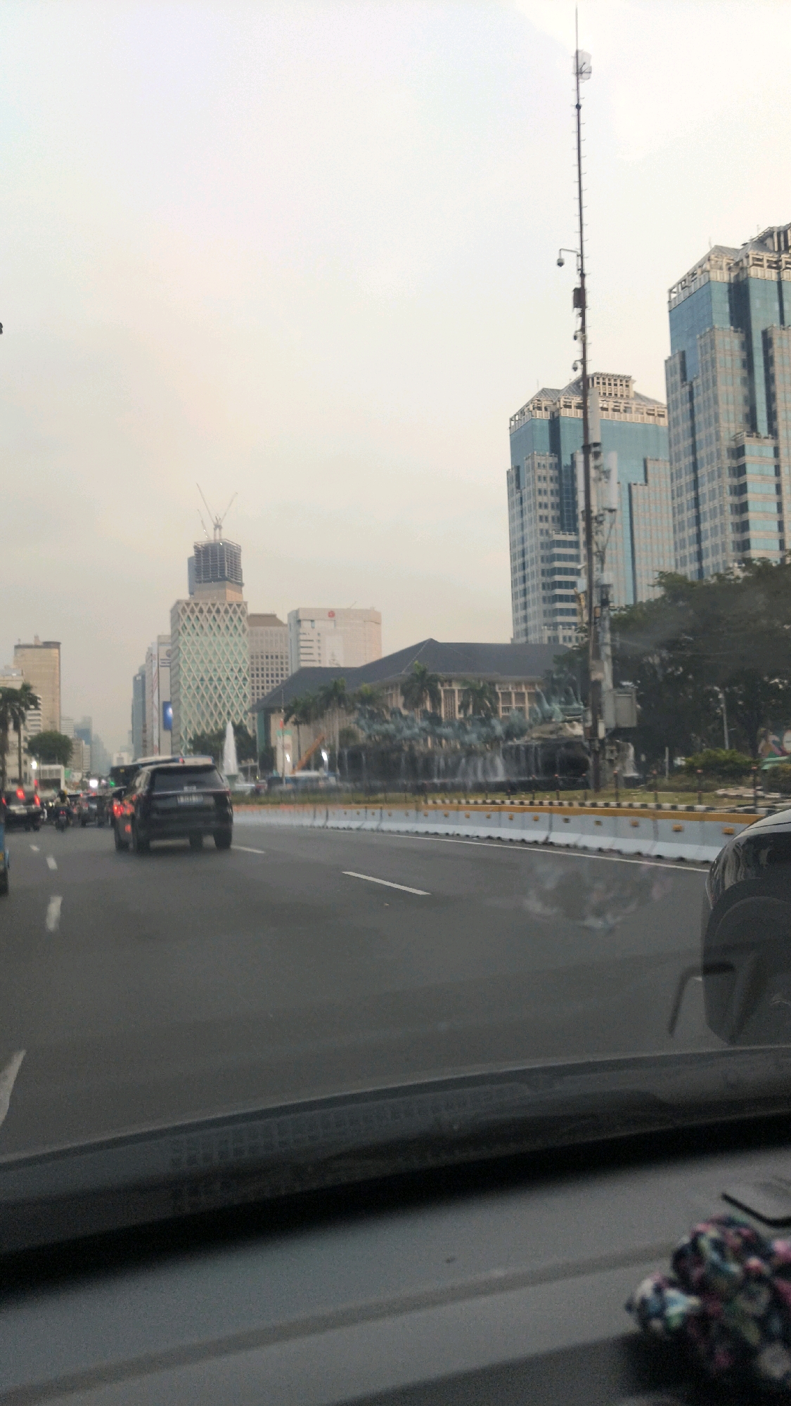 The image depicts a view from a vehicle traveling along a multi-lane road in the city of Central Jakarta, Indonesia, as evidenced by the architecture and the presence of a Jakarta Police car, though its markings are not clearly visible. The road is lined with traffic barriers. On the right side of the road, there is a large building with a dark, sloped roof adorned with sculpted figures and water features, reminiscent of a fountain or an artistic installation. Behind this building, tall, modern skyscrapers, characterized by their glass facades and angular designs, dominate the skyline. One particularly tall skyscraper, the Wisma 46 building, is visible to the right. A prominent telecommunications tower, equipped with multiple cameras and antennas, stands between the road and the skyscrapers. The sky is overcast with a pale, hazy light, suggesting either early morning, late afternoon, or a generally polluted atmosphere. Several vehicles, including cars and motorcycles, are present on the road, indicating moderate traffic. Construction cranes are also visible in the distance, signifying ongoing development in the area. The foreground of the image shows the interior of the vehicle, including the dashboard and a portion of the windshield.