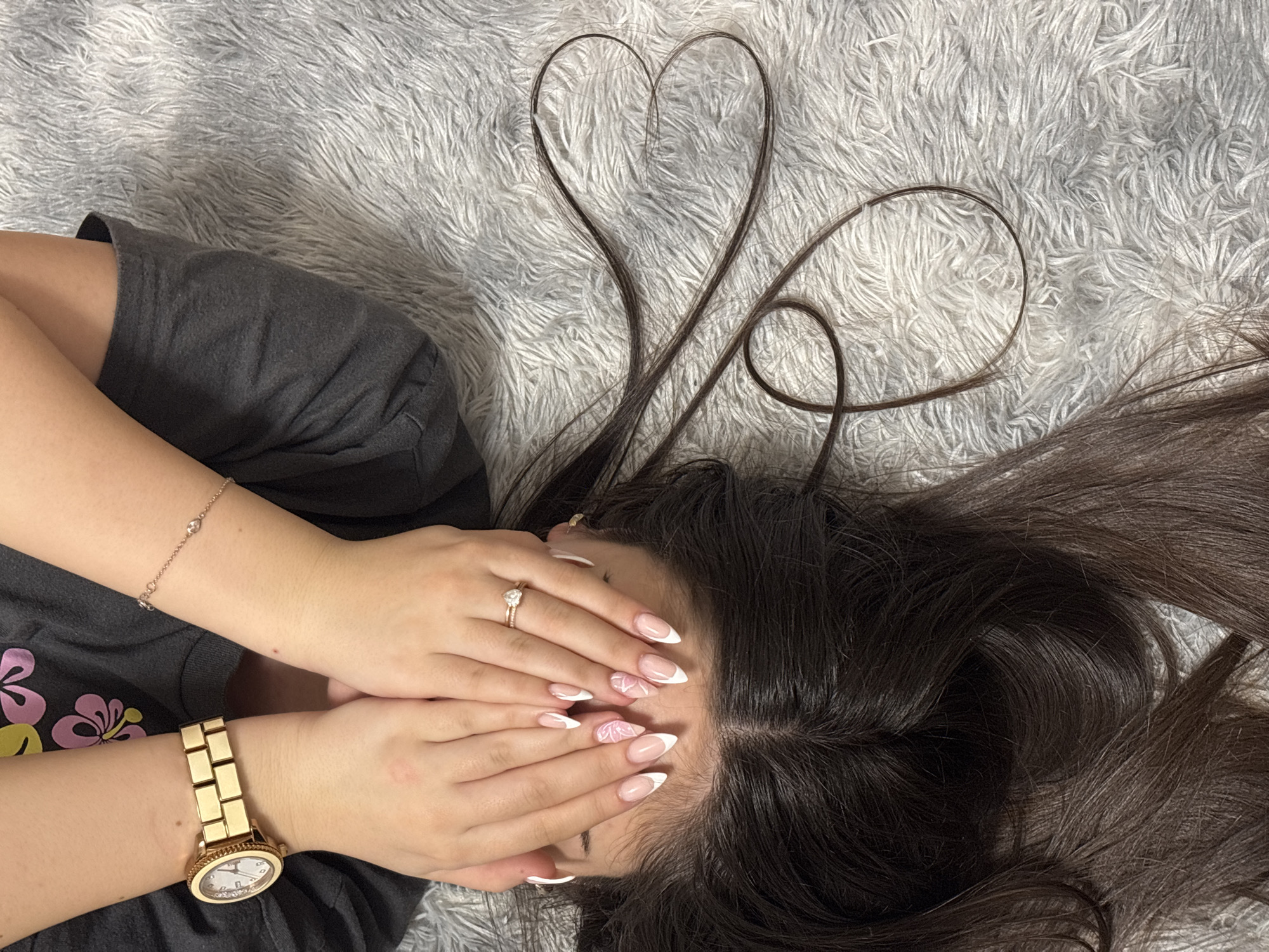 A person lies on a gray fuzzy rug with their hands covering their face. The person's dark brown hair is spread around their head on the rug. Two strands of hair are arranged to form a heart shape and a loop. The person is wearing a gray t-shirt with a pink and yellow flower design. On their left wrist is a gold watch, and on their right wrist is a rose gold bracelet. The person is wearing a ring on their left ring finger. Their fingernails are manicured with a white and pink design. The location is not determinable from this image, so Lechchevo, Bulgaria cannot be confirmed.