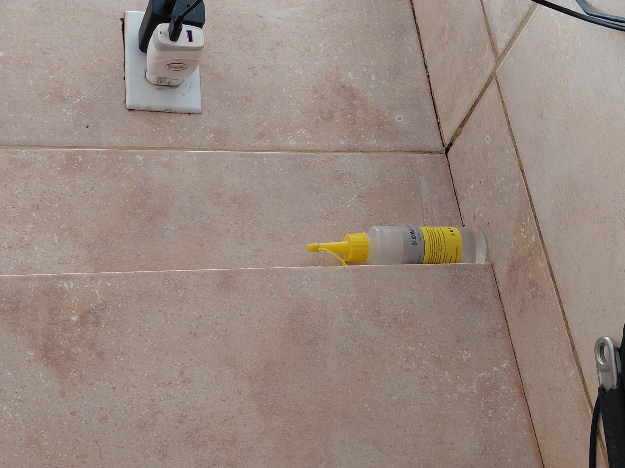 The image displays an indoor tiled corner featuring a power outlet, two plugged-in chargers, and a bottle of silicone sealant. The walls and a lower surface are covered in light beige-brown ceramic tiles with a speckled pattern. Grout lines are visible between the tiles, some appearing slightly discolored.
A white power outlet plate is affixed to a vertical tiled surface. Two USB wall chargers are plugged into it; one is black and mostly obscured, while the other is white with a blue USB port. On a lower horizontal tiled surface, a clear plastic bottle of silicone sealant with a yellow nozzle and cap lies on its side. Part of a black strap with a silver metal hook is visible on the far right edge of the frame.
There is no specific activity taking place; the scene captures a static arrangement of objects in what appears to be a functional or utility area. The lighting suggests an indoor setting.
Visible text includes "Dowy" and a "CE" marking on the white charger. On the silicone sealant bottle, "SILICONA" and "LÍQUIDAS" are clearly legible, along with the abbreviation "ES". Other text on the yellow label of the bottle is too small to decipher.