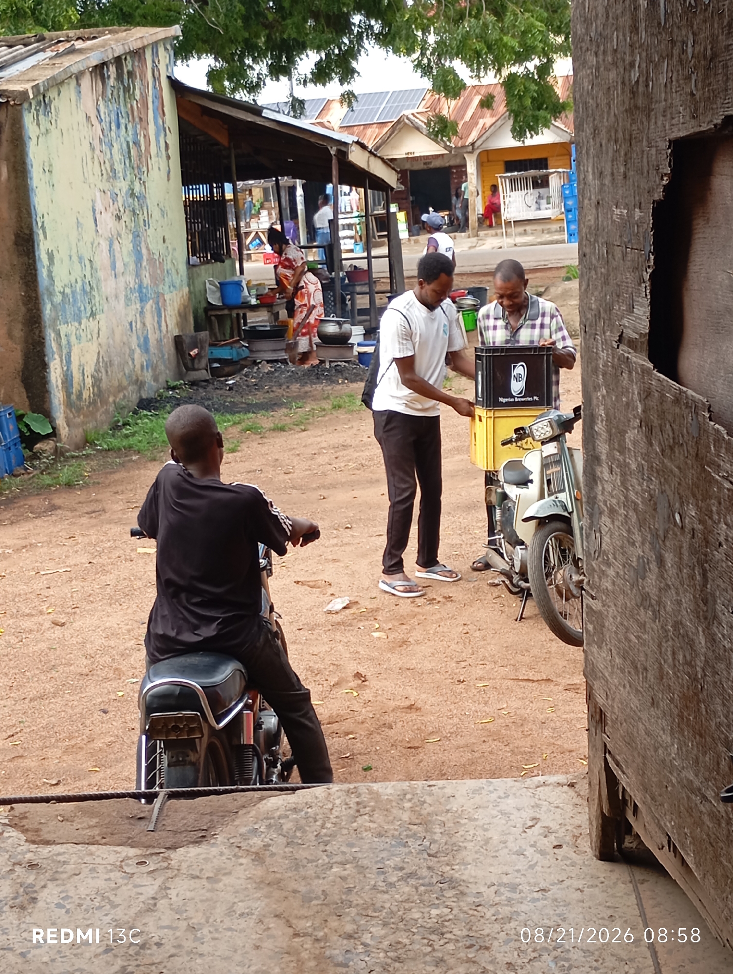 The image depicts an outdoor scene in the city of Shendam, Nigeria, where two men are interacting near a parked motorcycle. One man, wearing a white t-shirt and black pants, is extending his hand towards a yellow crate. The other man, dressed in a plaid shirt and dark pants, is holding a black crate labeled "NB Nigerian Breweries Plc." The motorcycle, a light-colored scooter, is parked on a dirt path. In the background, a woman in a patterned dress is visible near a stall with cooking equipment, suggesting a market or roadside vendor area. The environment is somewhat rustic, with a dilapidated building on the left and a wooden structure on the right. The ground is covered with dirt and scattered leaves. The timestamp in the lower right corner indicates the image was captured on August 21, 2026, at 08:58.
